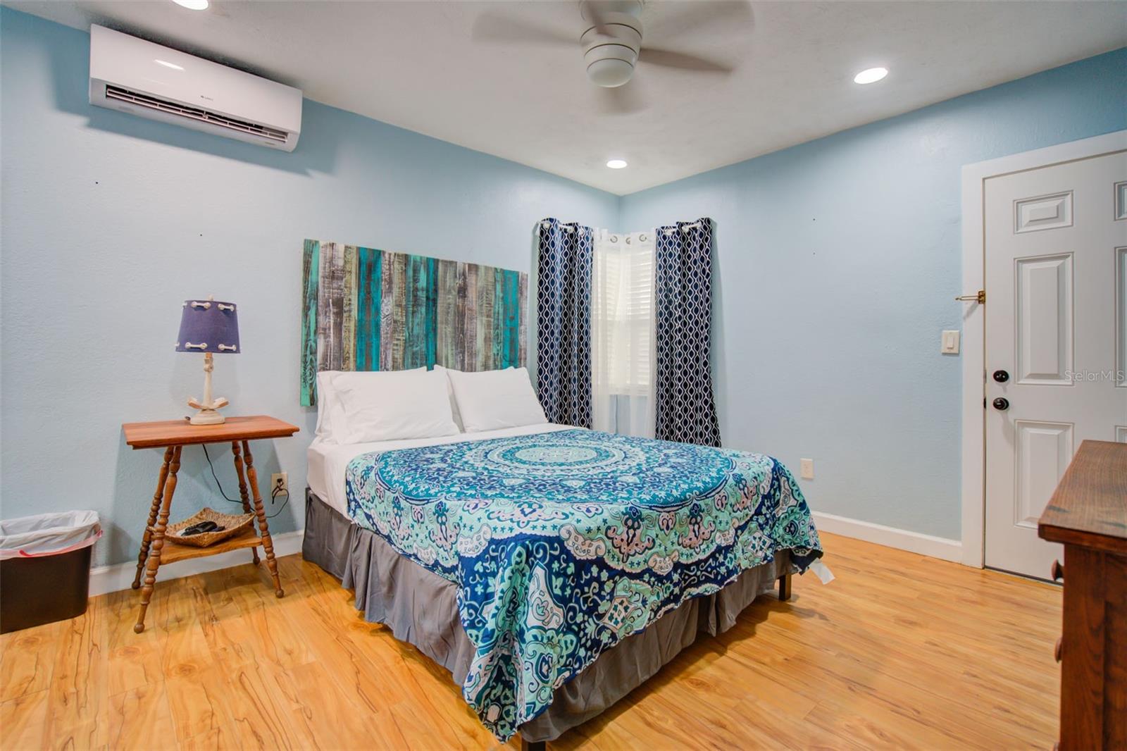 Furnished bedroom with a blue patterned bedspread, light blue walls, and wood flooring.
