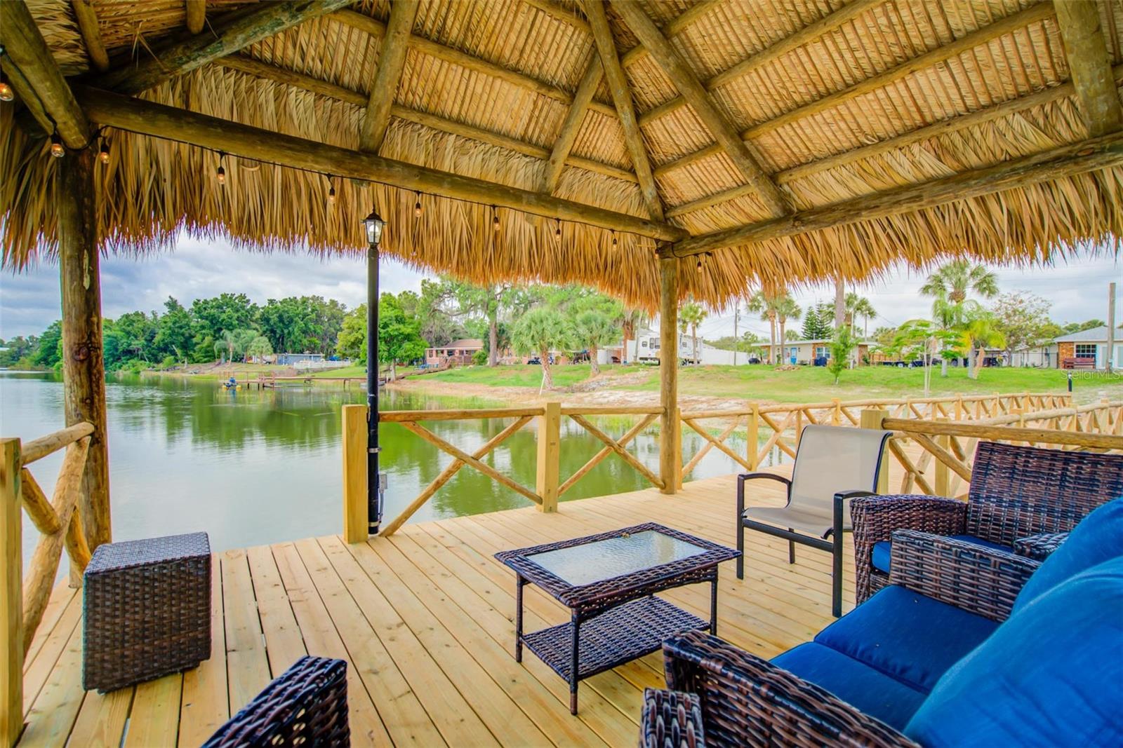 Furnished waterfront deck beneath a thatched tiki roof, with wicker seating and views across the water.