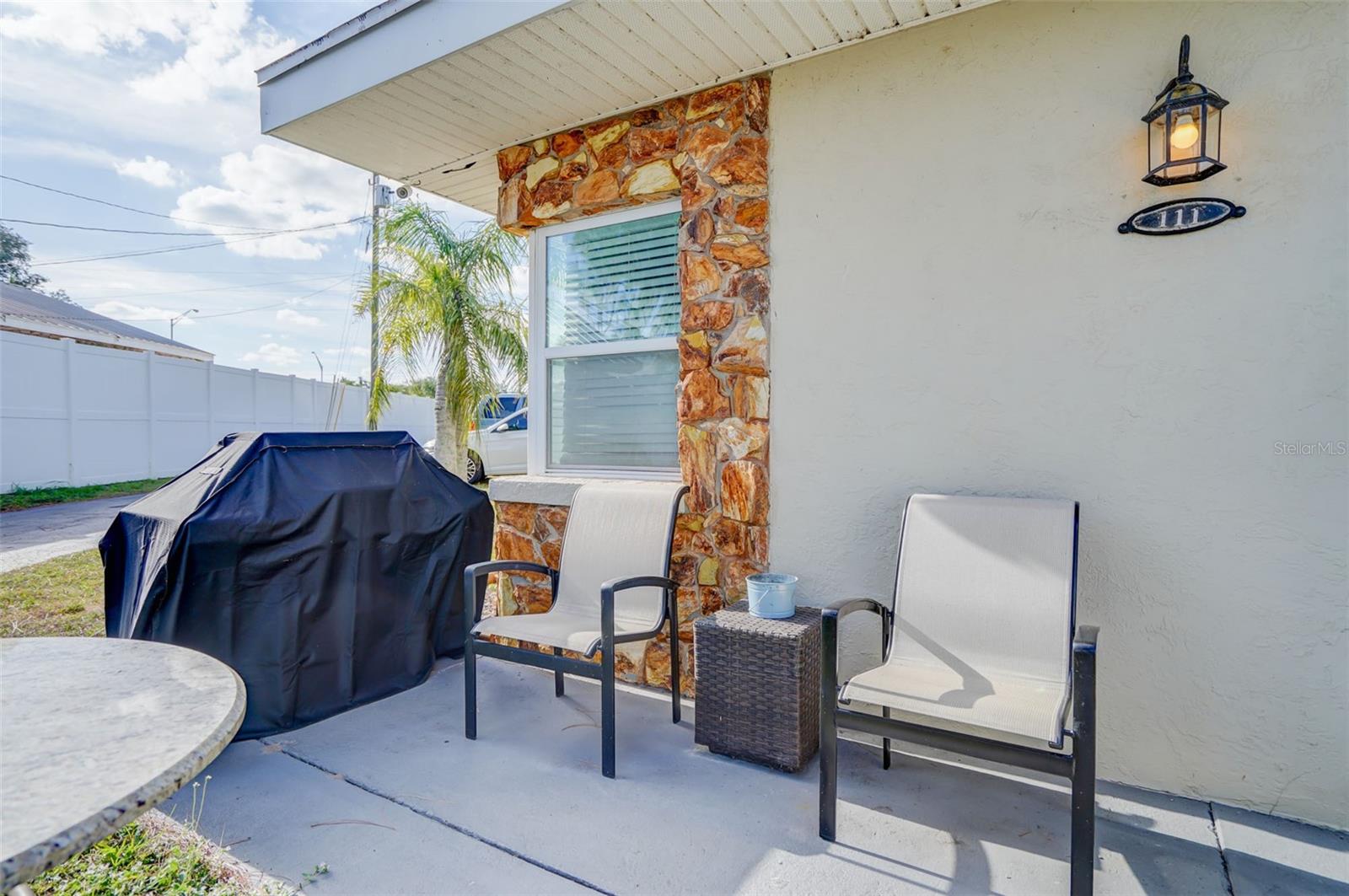 Covered patio with two chairs and a covered grill beside a stone-trimmed window.