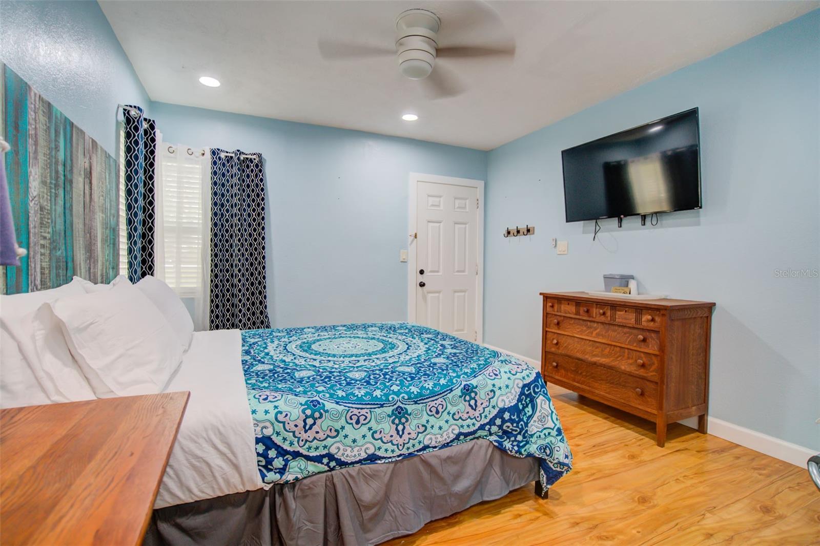 Furnished bedroom with blue patterned bedding, a wooden dresser, and a wall-mounted television.