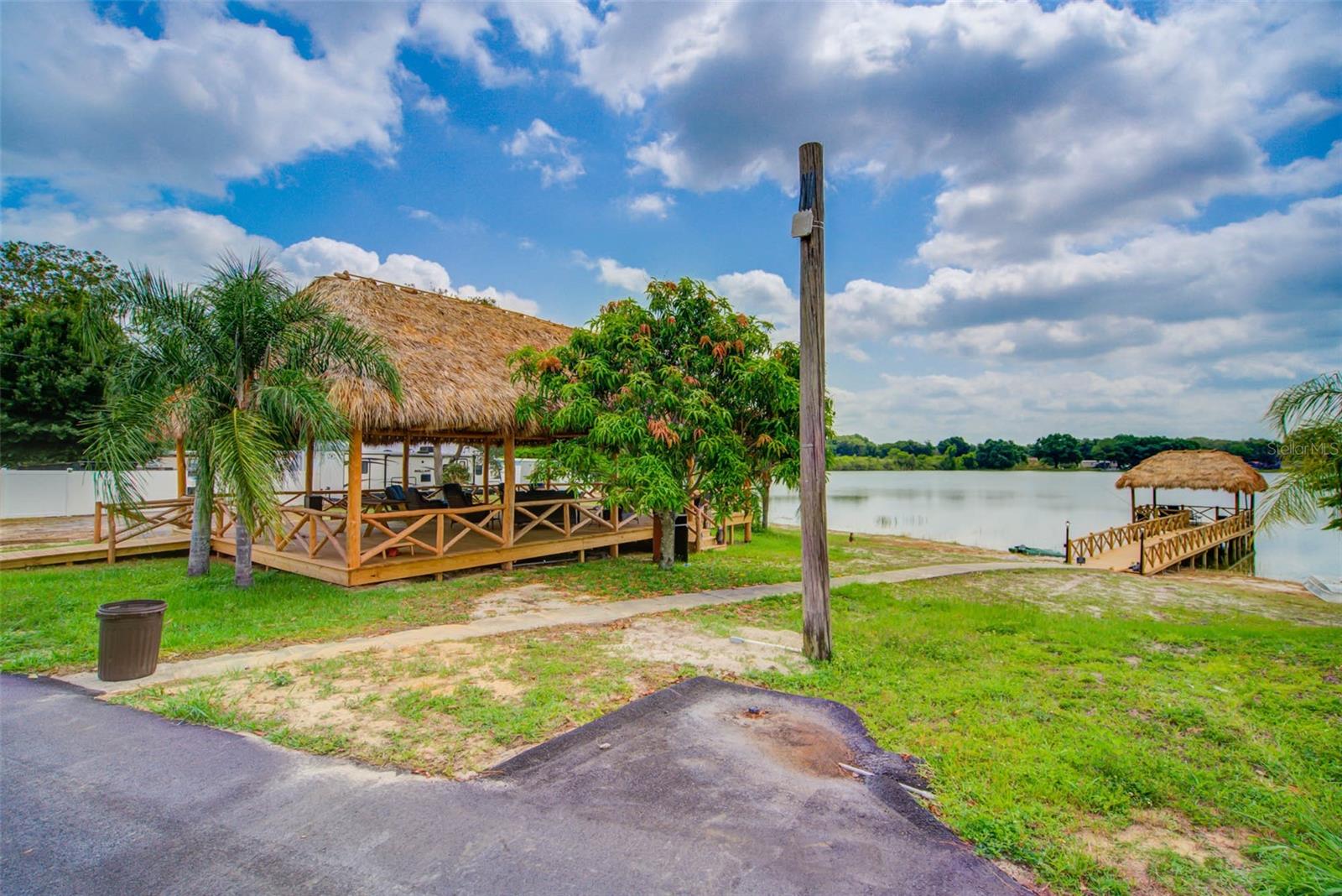 Waterfront pavilion and wooden dock beside a calm lake, surrounded by palms and lawn.