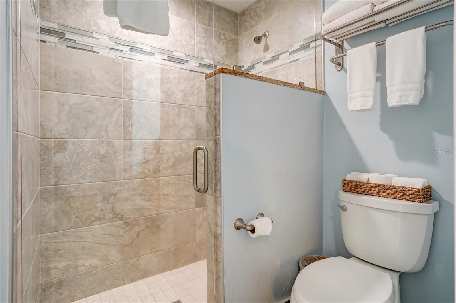 Bathroom with a tiled glass shower and white toilet topped by a basket of towels.