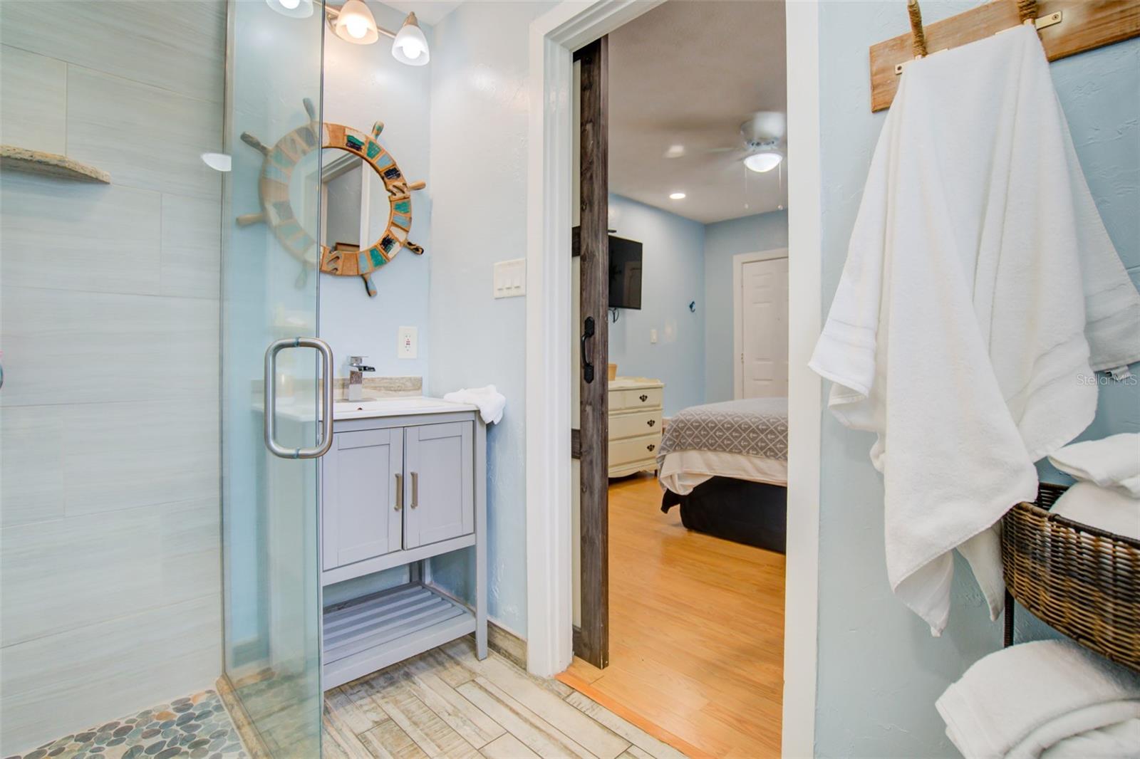 Bathroom with a glass shower, pebble-tile floor, light vanity, and nautical mirror beside a bedroom doorway.