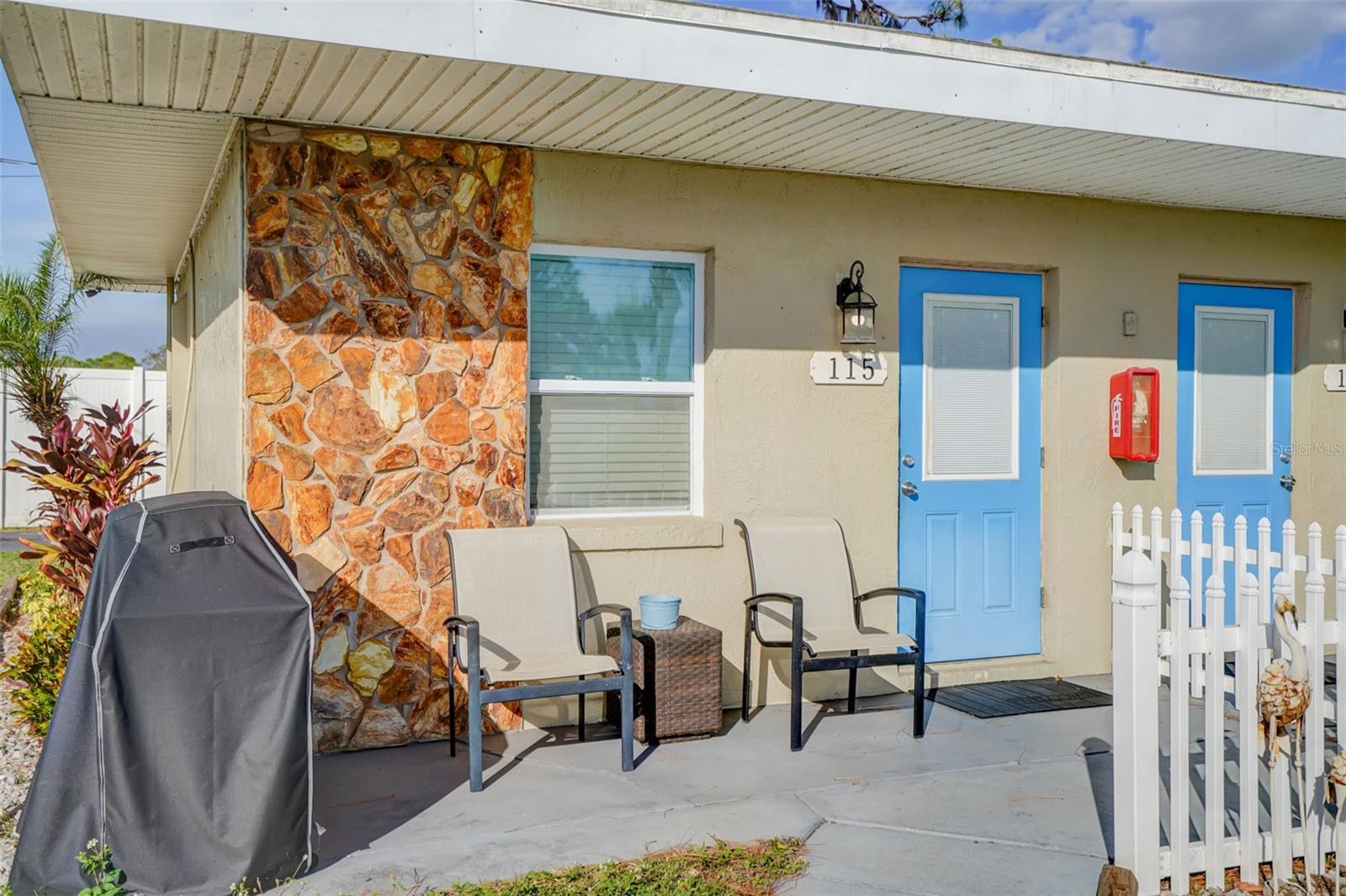 Covered patio with stone accent wall, blue entrance door, outdoor chairs, and white picket fence.
