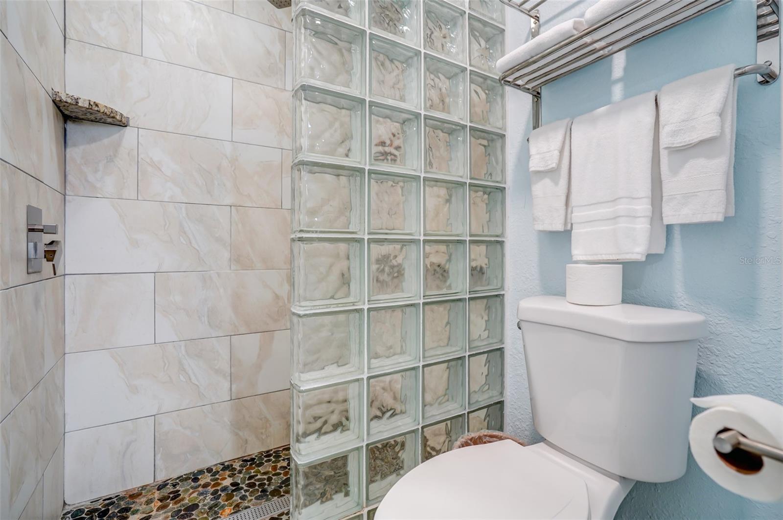 Bathroom shower with glass-block partition and pebble-stone floor beside a toilet.