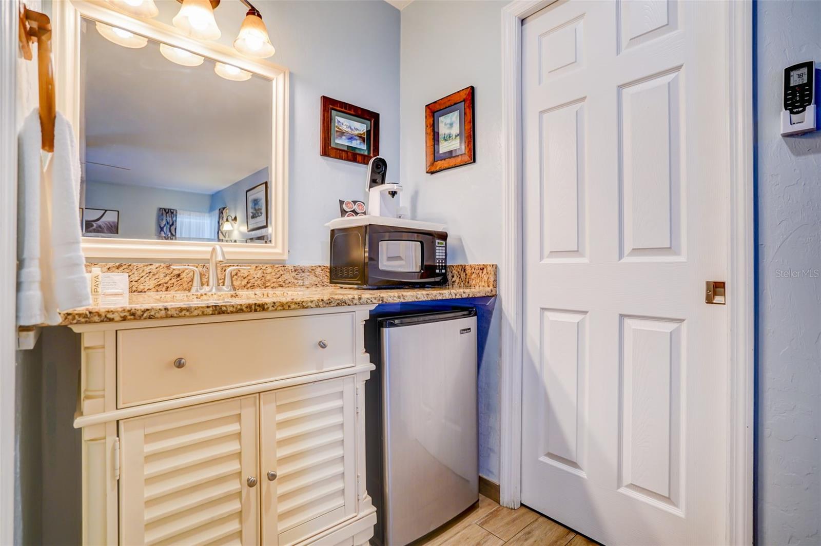 Bathroom vanity with granite counter, large mirror, microwave, and compact stainless-steel refrigerator.