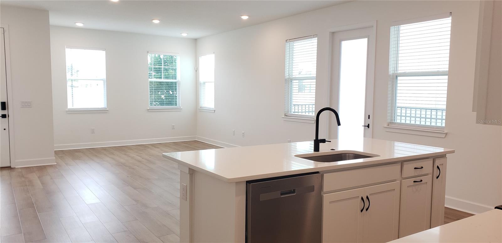 Open kitchen with white island, black faucet, stainless dishwasher, and bright adjoining living area.
