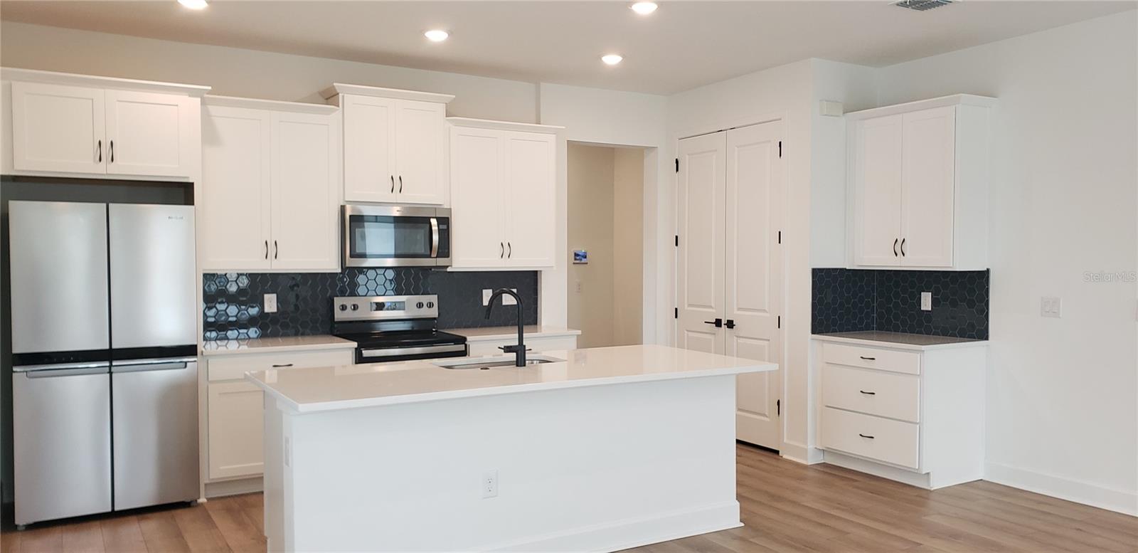 Kitchen with a large white island, stainless steel appliances, and dark hexagonal tile backsplash.