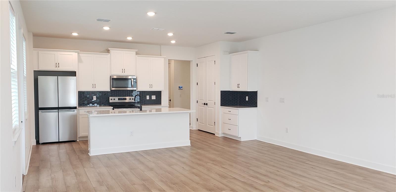 Open kitchen with white cabinets, dark hexagonal backsplash, stainless appliances, and a wide island.