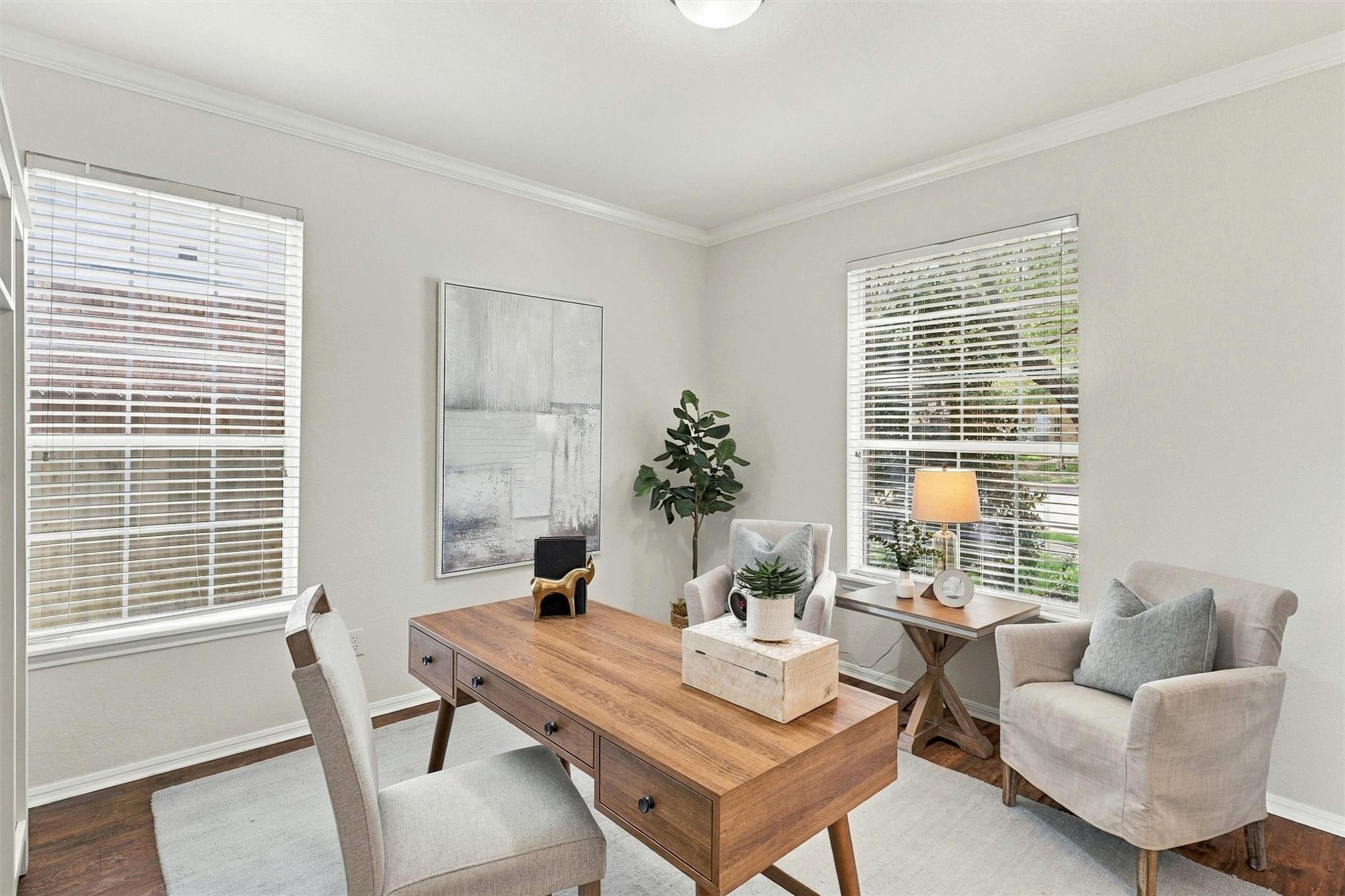 Staged home office with a large wooden desk and upholstered seating beside two windows.