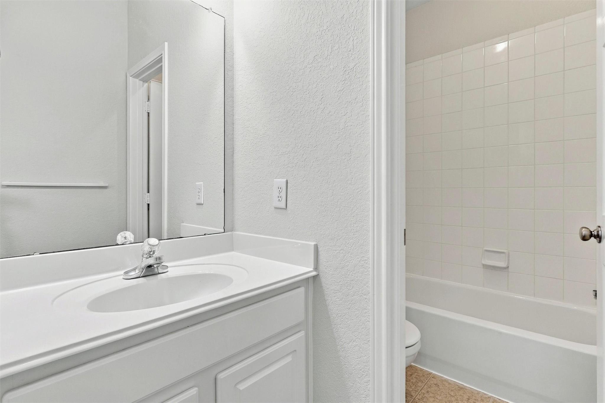 Bathroom with a white vanity and mirror beside a bathtub with tiled walls.