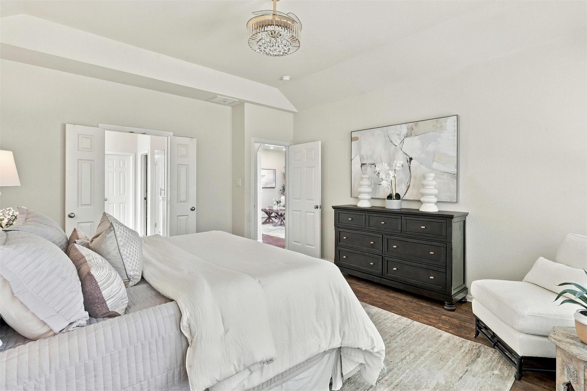 Furnished bedroom with a white bed, dark dresser, chandelier, and open doors to adjoining rooms.