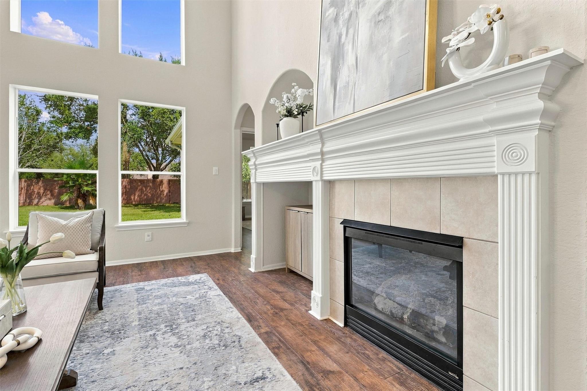 Living room with a large tiled fireplace, ornate white mantel, and tall windows overlooking a fenced yard.