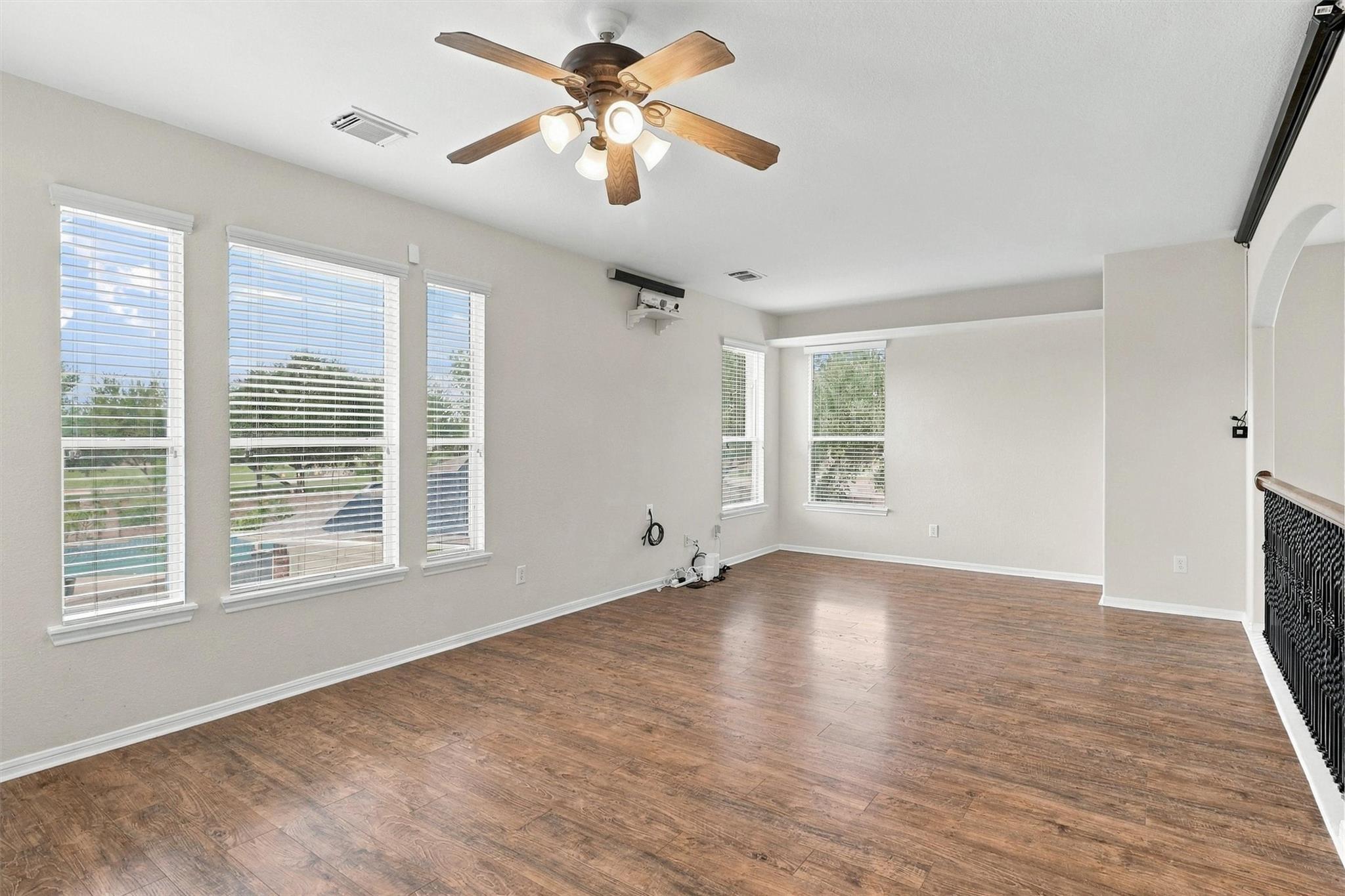 Empty living area with wood-look flooring, multiple windows, ceiling fan, and wall-mounted projector.