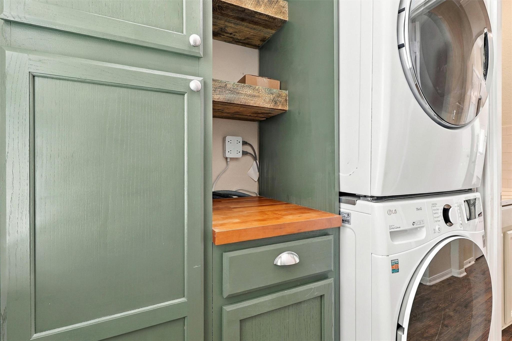Laundry room with stacked white washer and dryer beside sage green cabinets and a wood countertop.