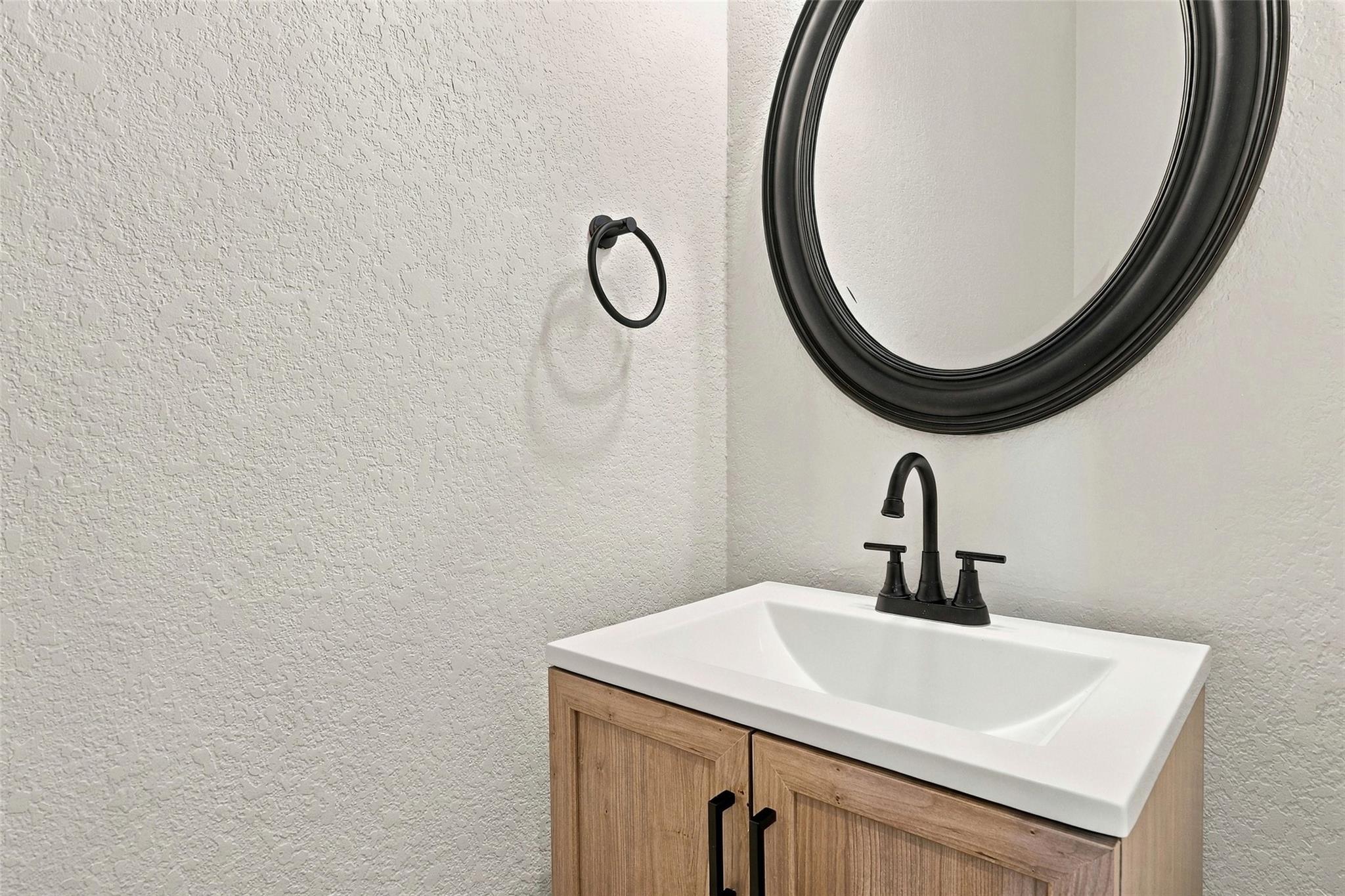 Bathroom vanity with a white sink, light wood cabinet, and matte black fixtures beneath a round mirror.