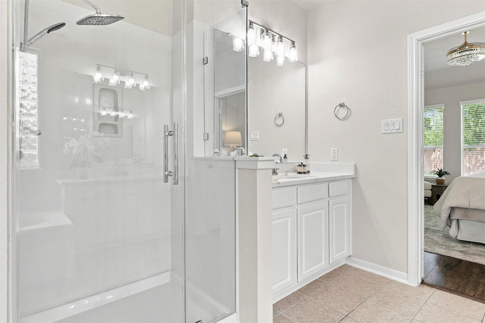 Bathroom with a glass-enclosed shower, mirrored vanity, white cabinetry, and tan tile floor.