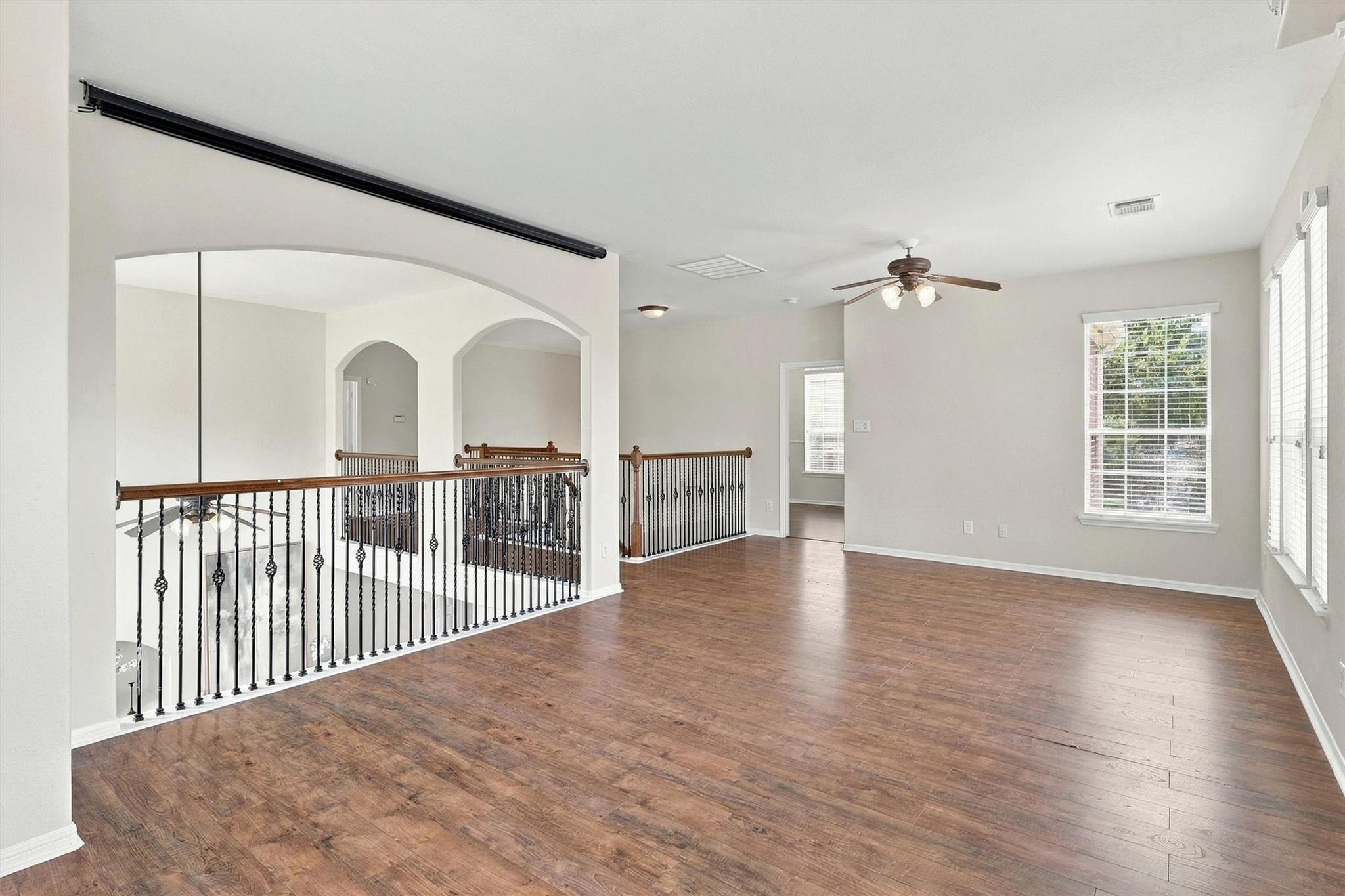 Empty upstairs loft with wood-look flooring, ceiling fan, and decorative iron railing overlooking the floor below.