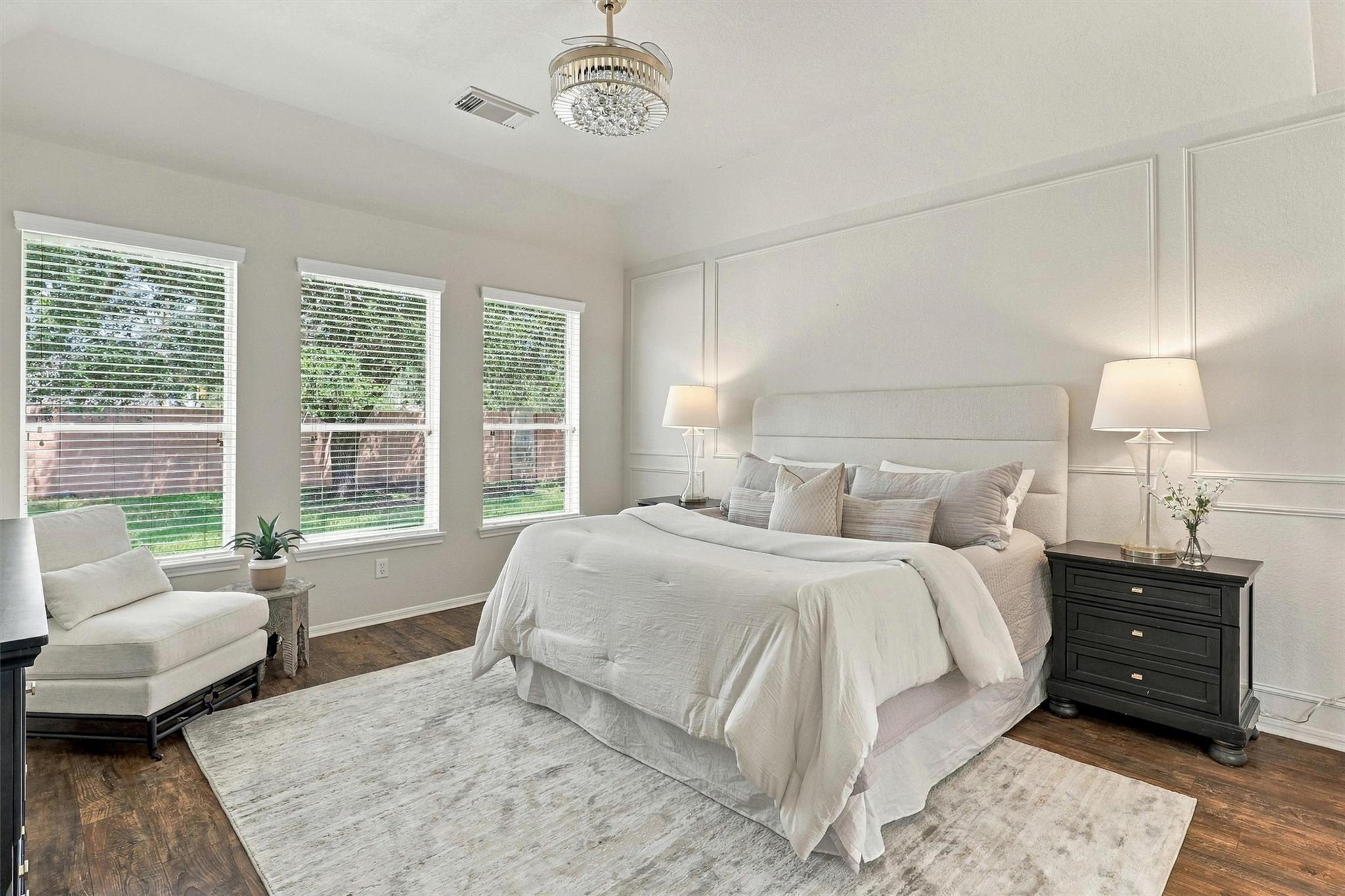 Bright bedroom with a large white bed, paneled walls, and three windows overlooking the yard.