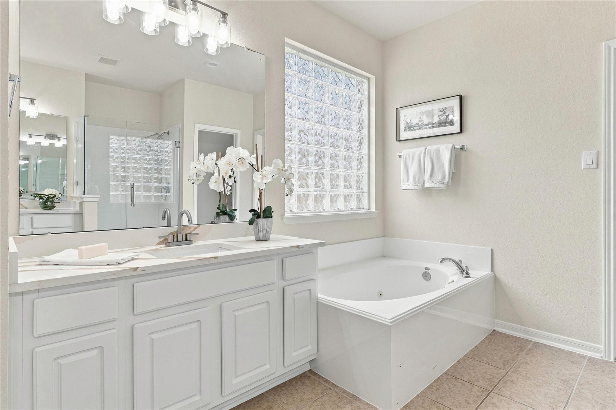 Bright bathroom with white vanity, glass-block window, corner soaking tub, and glass shower.