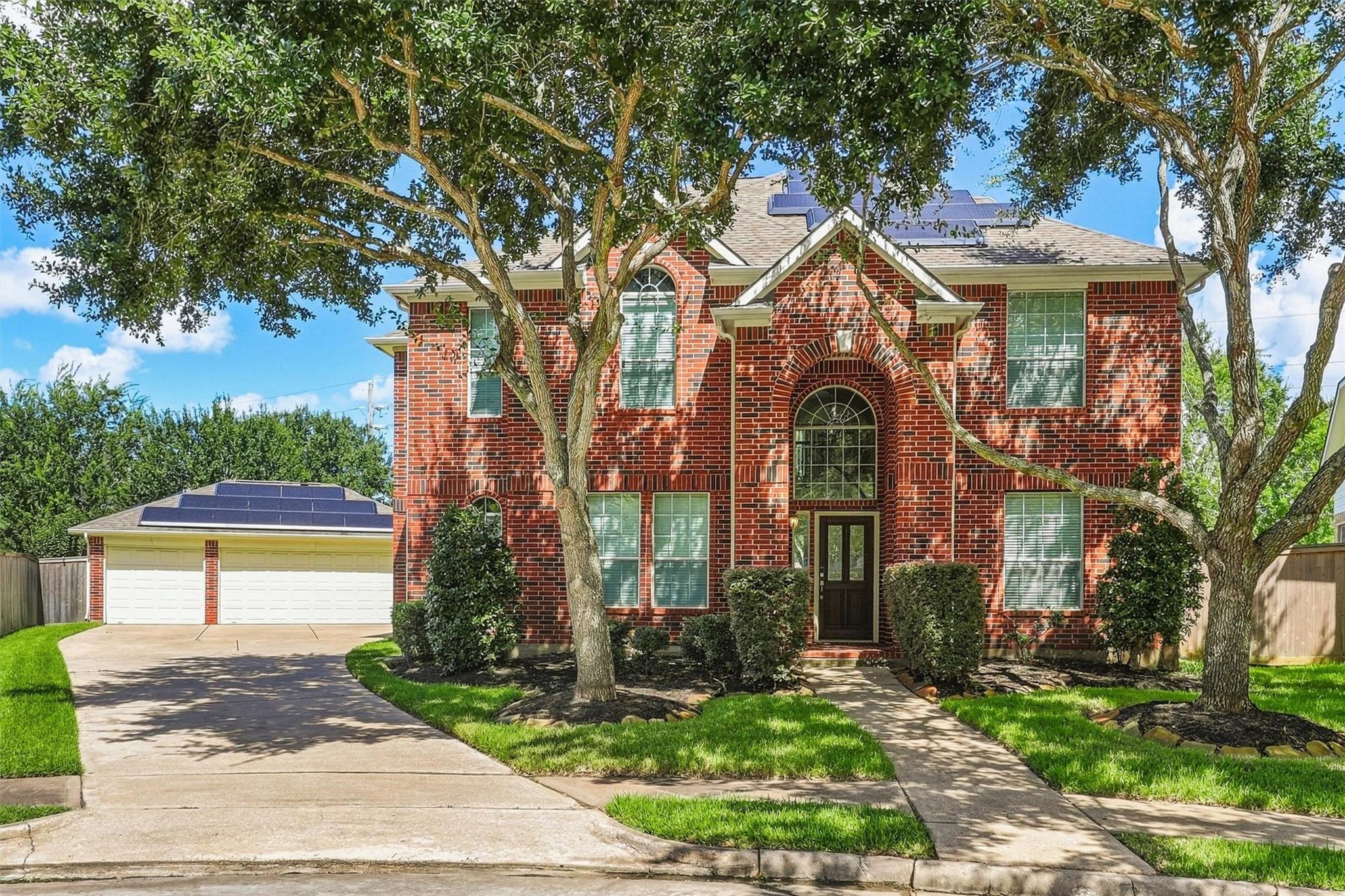 Two-story red-brick home with rooftop solar panels, a double garage, and mature shade trees.