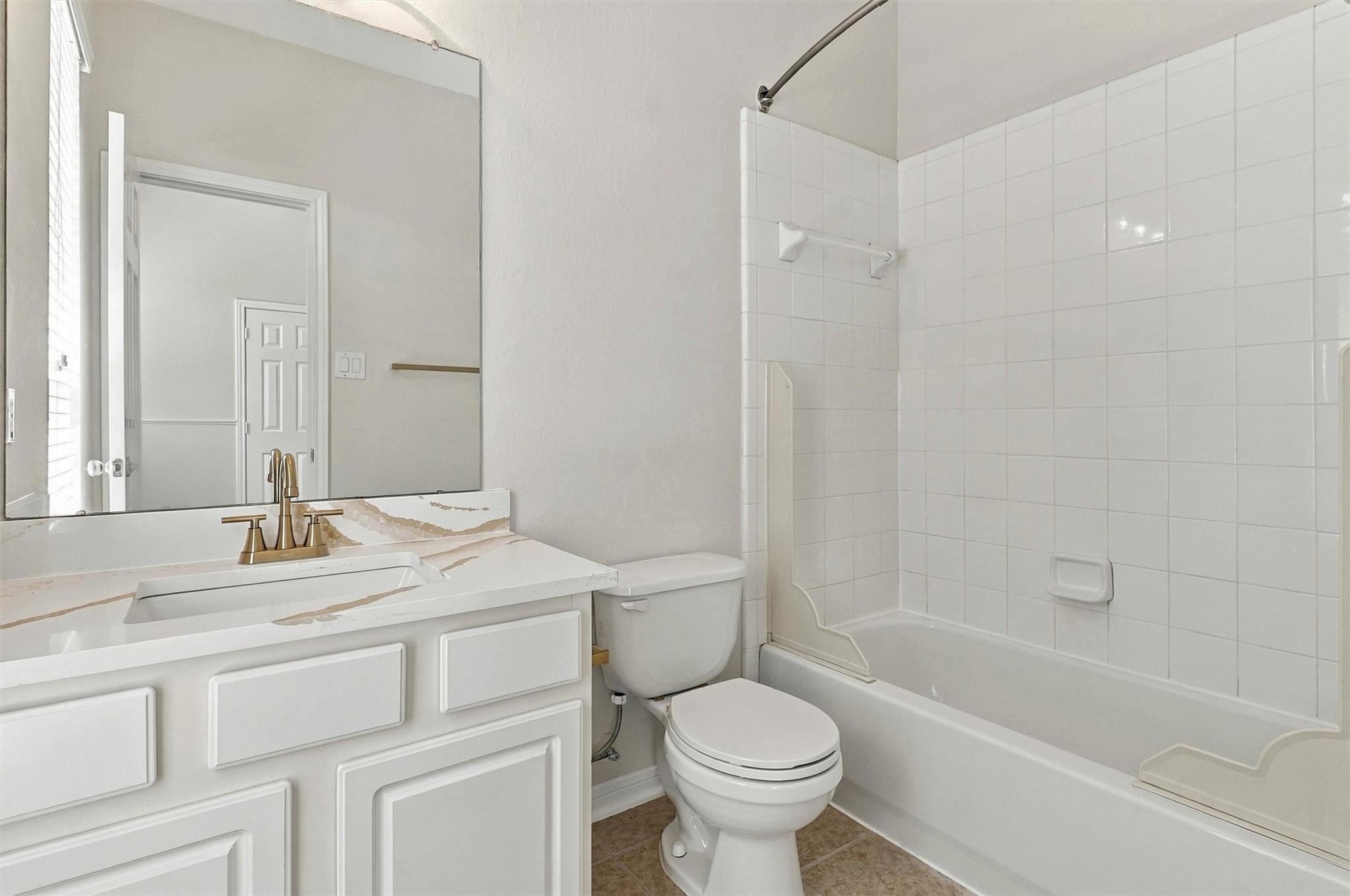 Bathroom with a marble-look vanity, brass faucet, toilet, and white tiled tub surround.