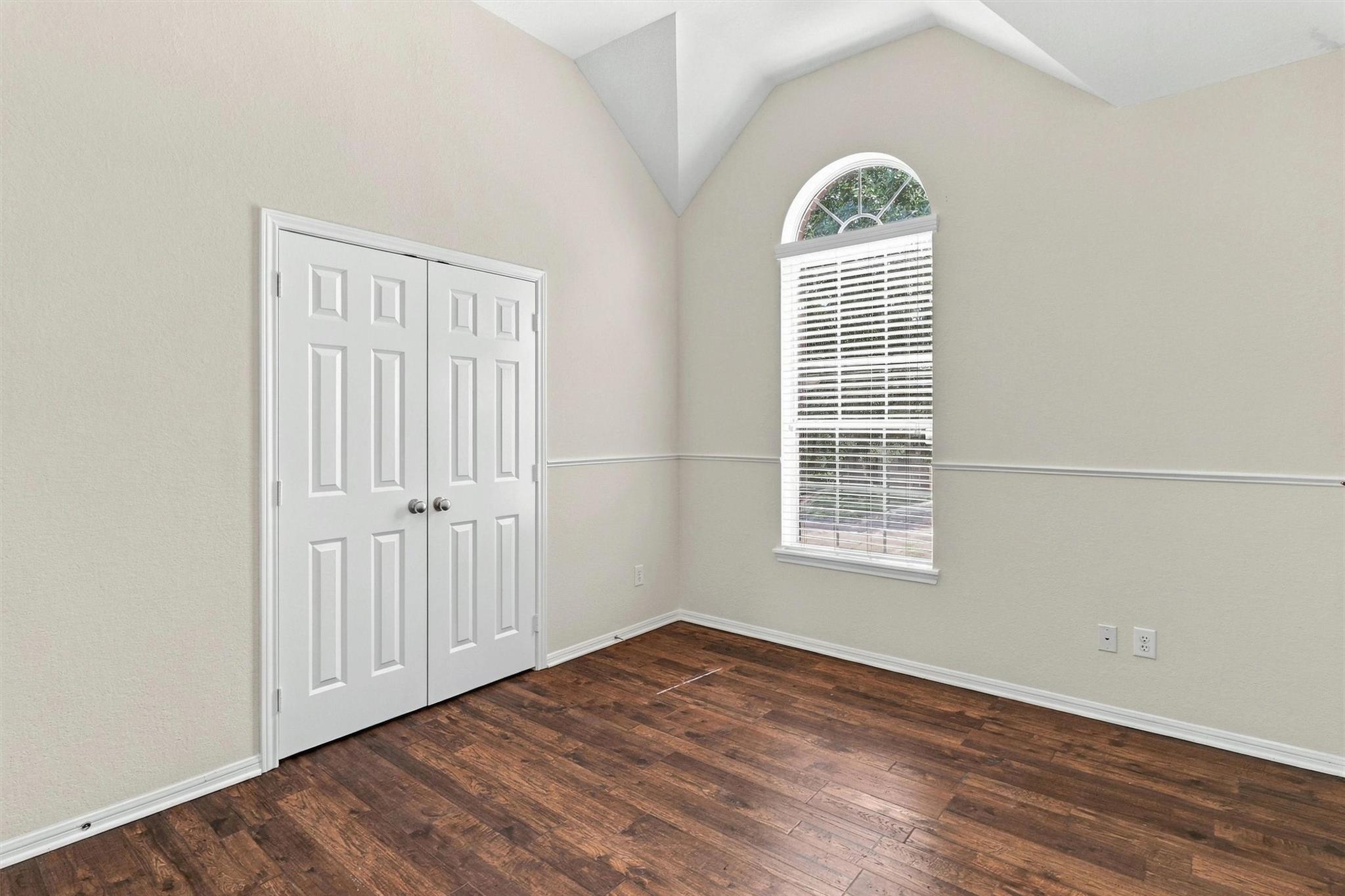 Empty room with dark wood floors, double closet doors, and a tall arched window.