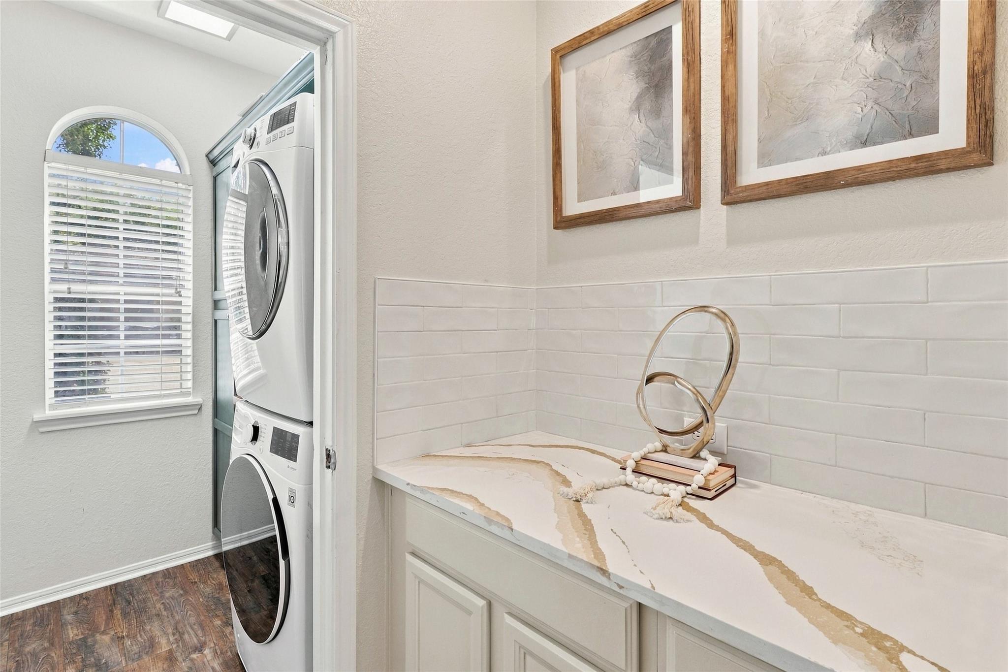 Laundry room with stacked washer and dryer beside a veined stone countertop and arched window.