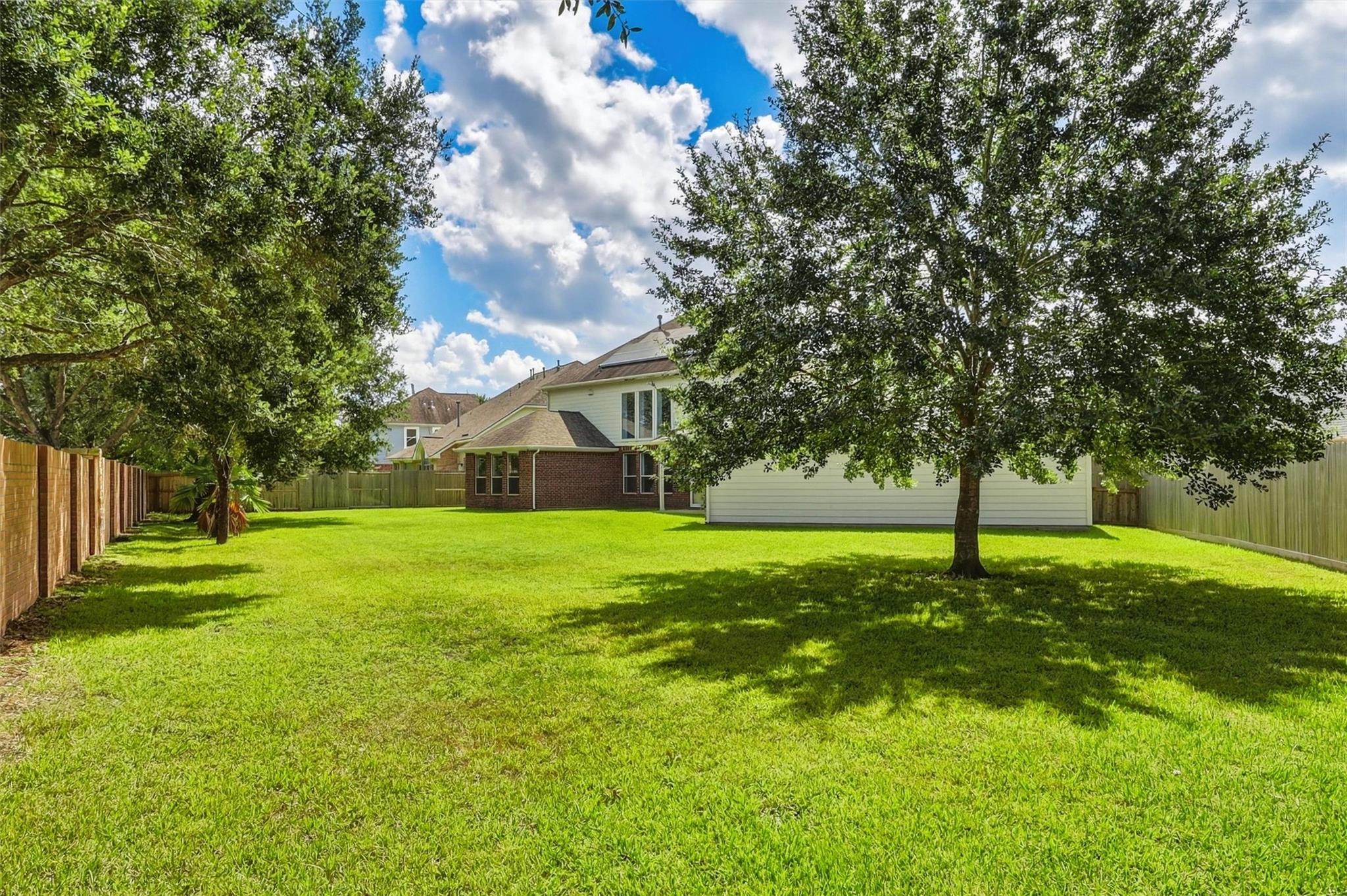 Spacious backyard with a broad lawn, mature shade trees, wood fencing, and the rear of a two-story home.