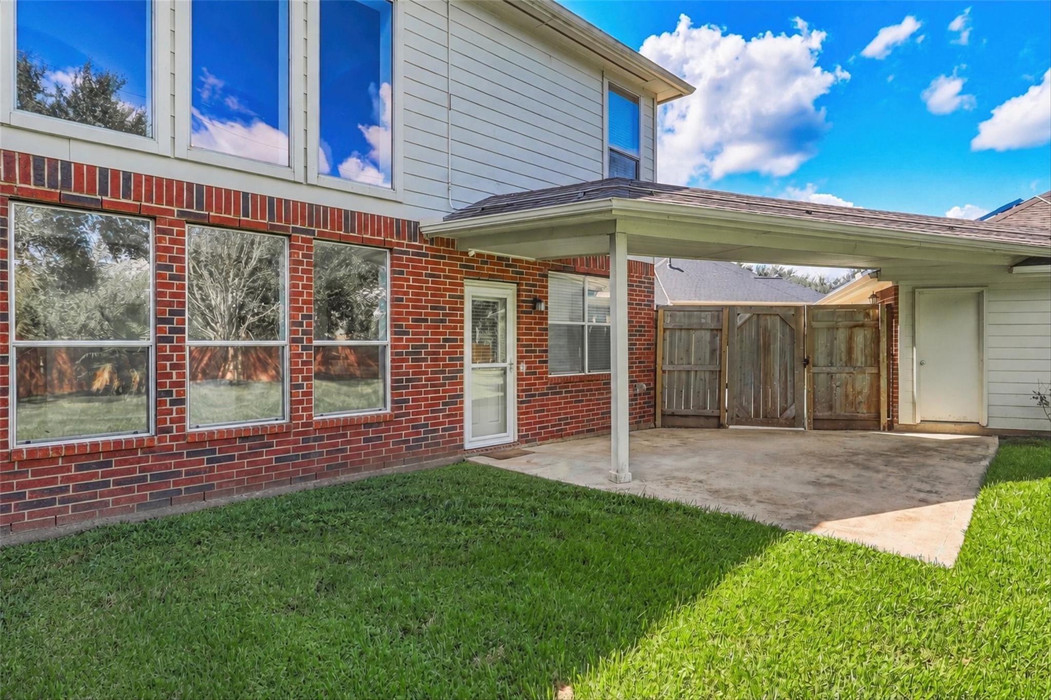 Rear of a two-story brick-and-siding home with a covered patio, wooden gate, and green lawn.