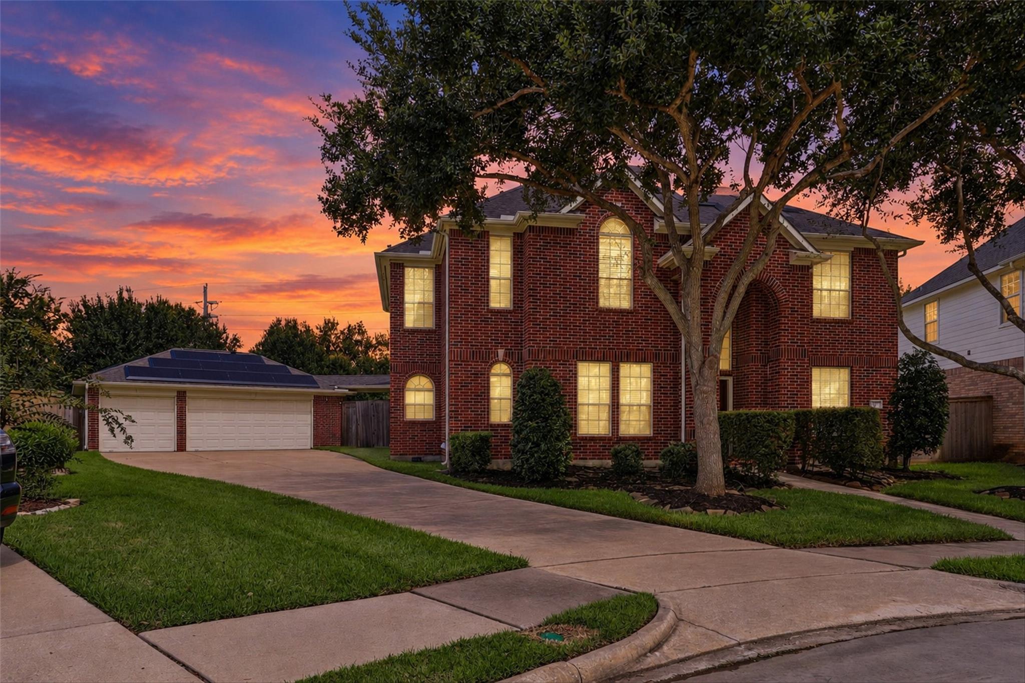 Two-story brick home with solar panels on the garage roof, glowing windows, and a large front-yard tree at dusk.