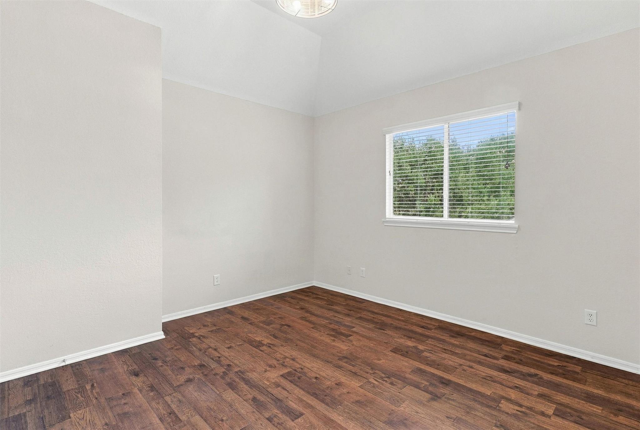 Empty room with dark wood floors and a window with blinds overlooking trees.