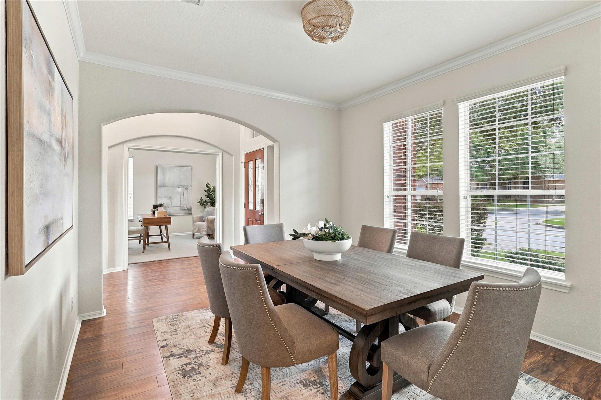 Dining room with a wooden table and upholstered chairs beside wide windows, opening into a sitting room.