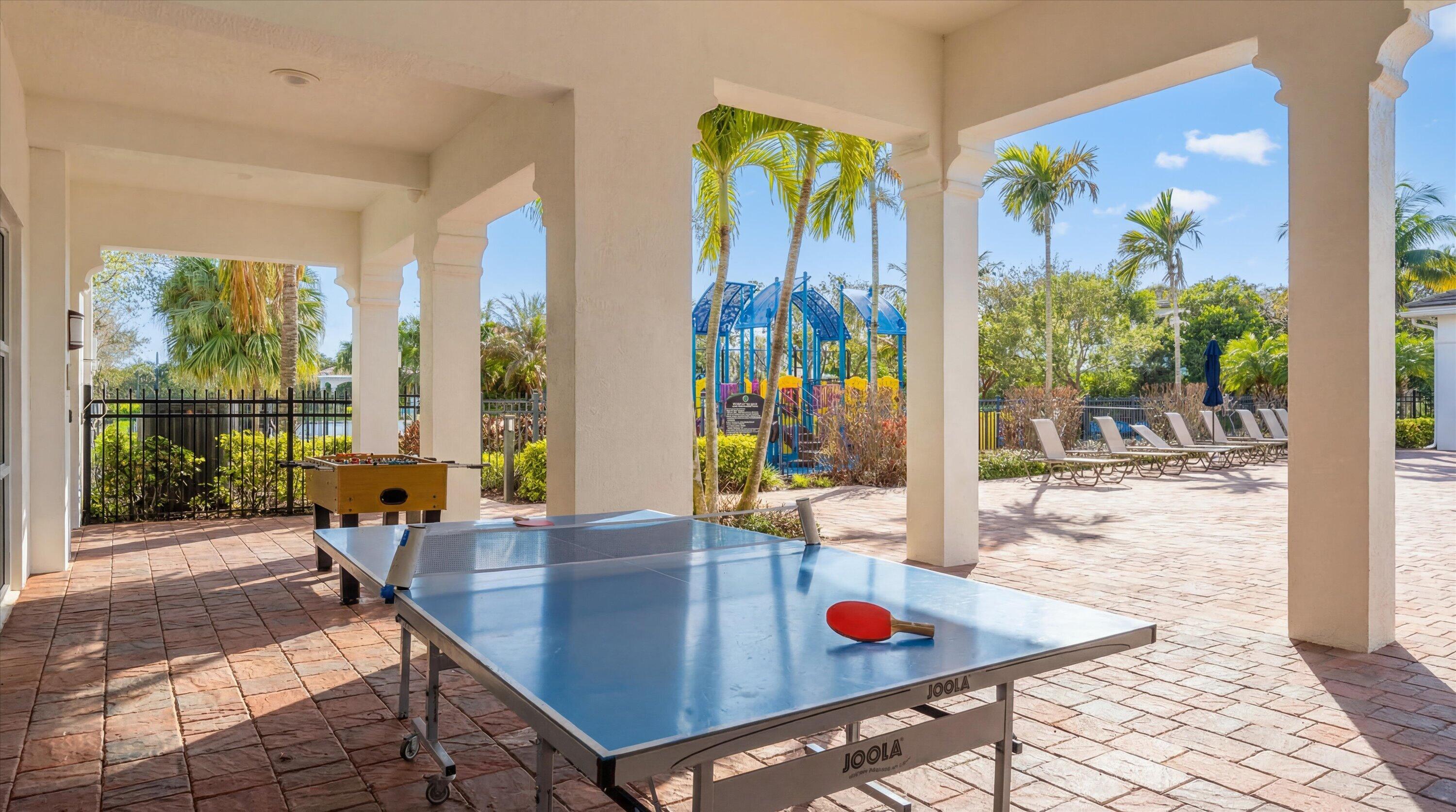 Covered recreation patio with ping-pong and foosball tables, a colorful playground, and palm trees.