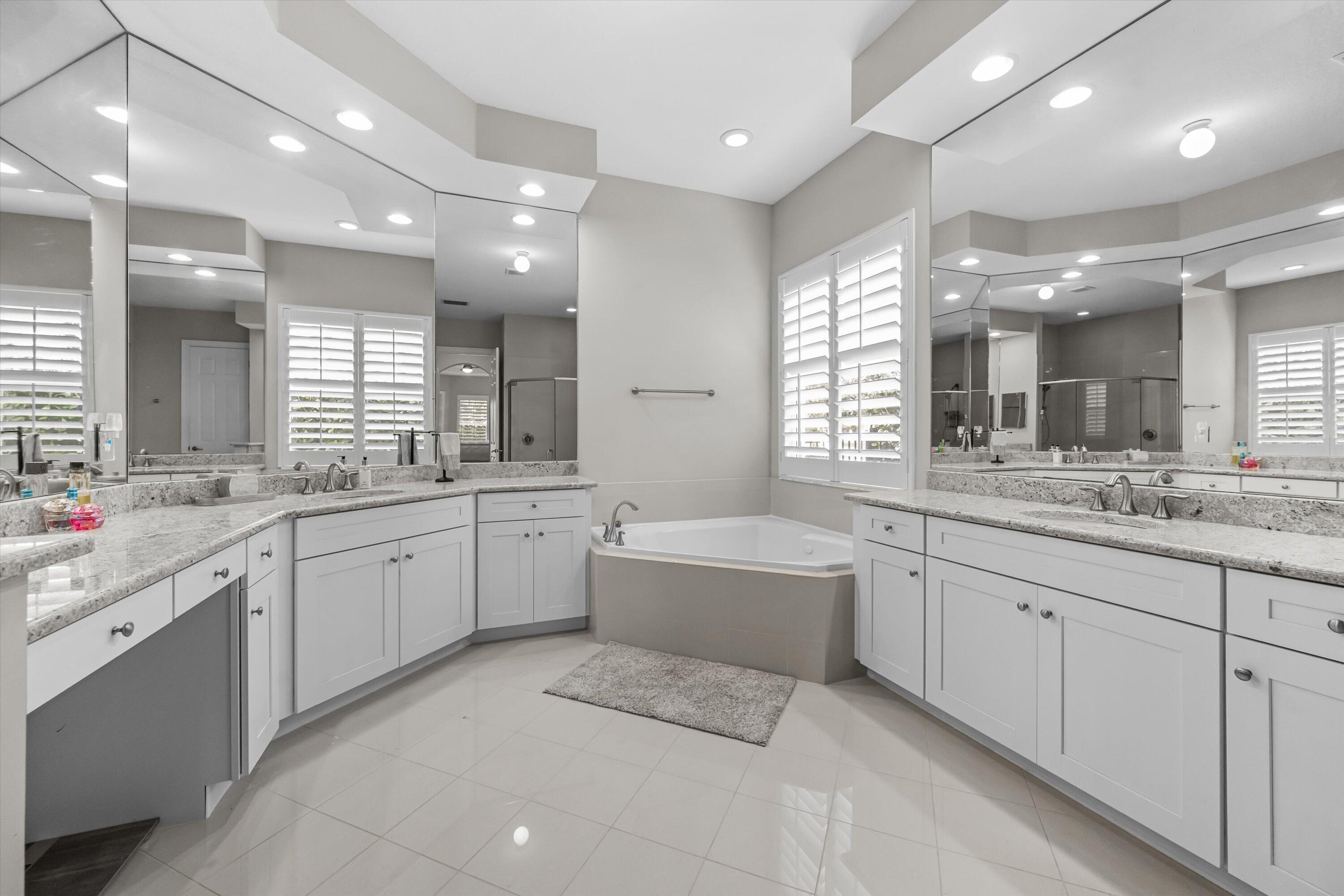 Primary bathroom with a built-in soaking tub, double vanities, granite counters, and shuttered windows.