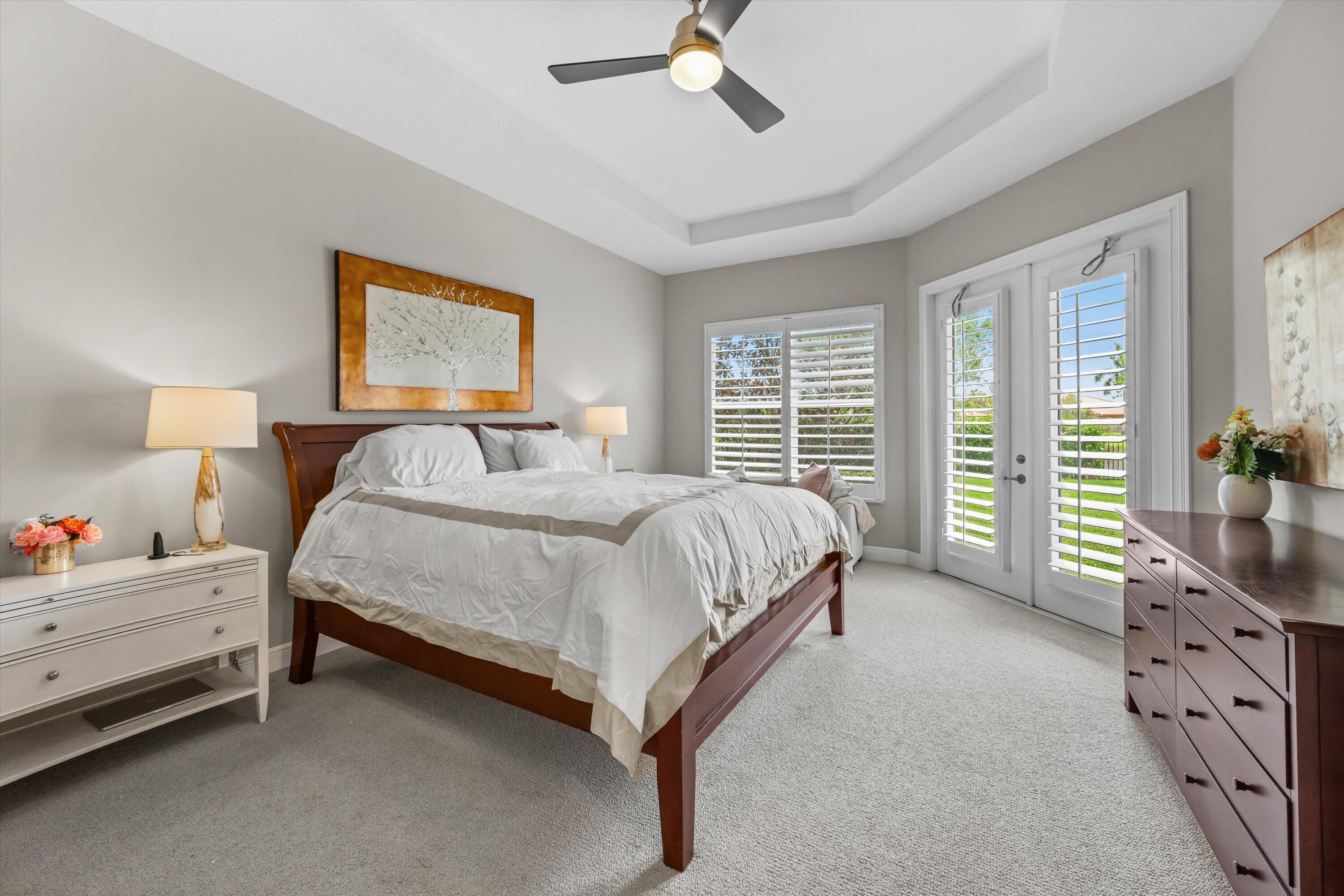 Primary bedroom with a dark wood bed, tray ceiling, and shuttered French doors opening to the lawn.
