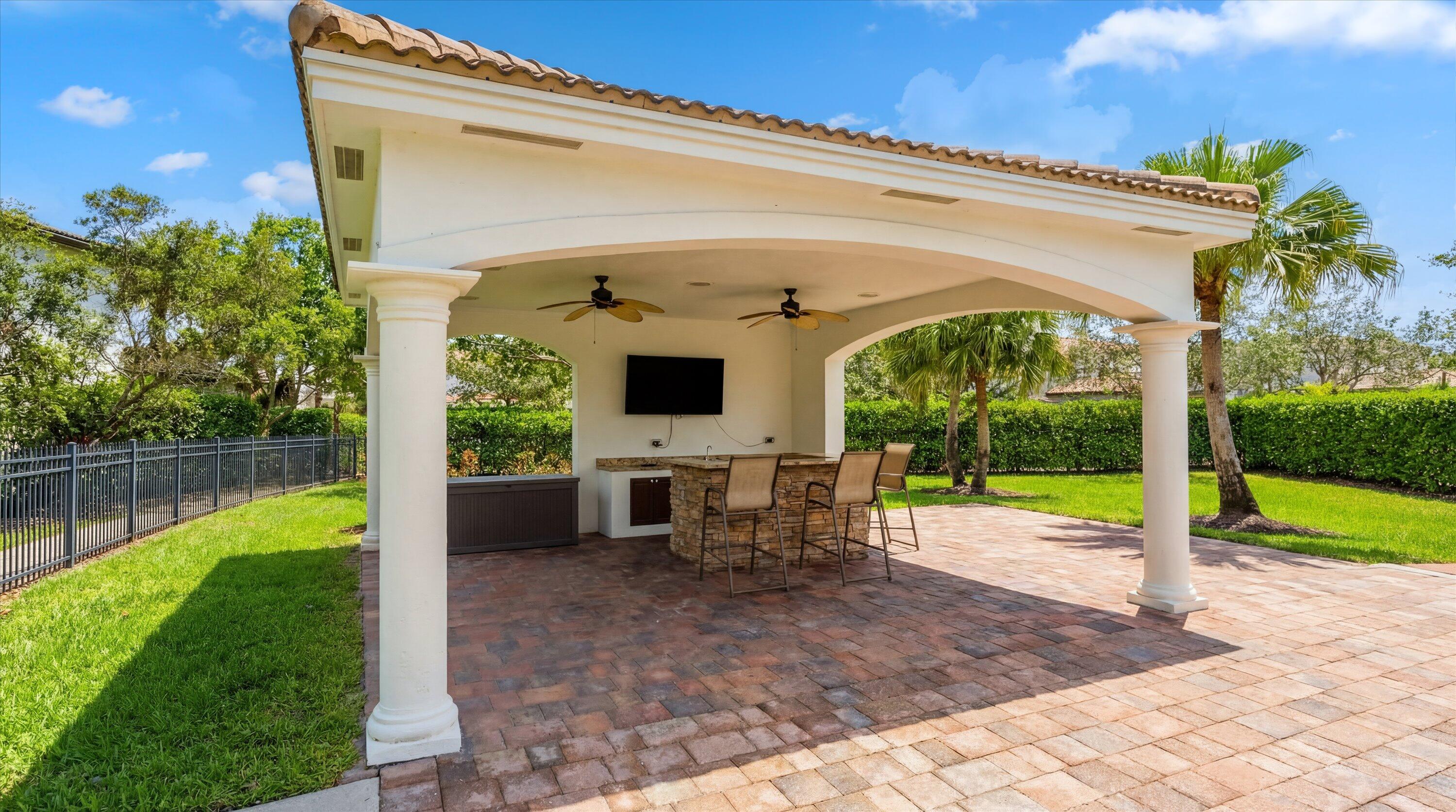 Covered patio with a stone outdoor kitchen, bar stools, ceiling fans, and brick pavers.