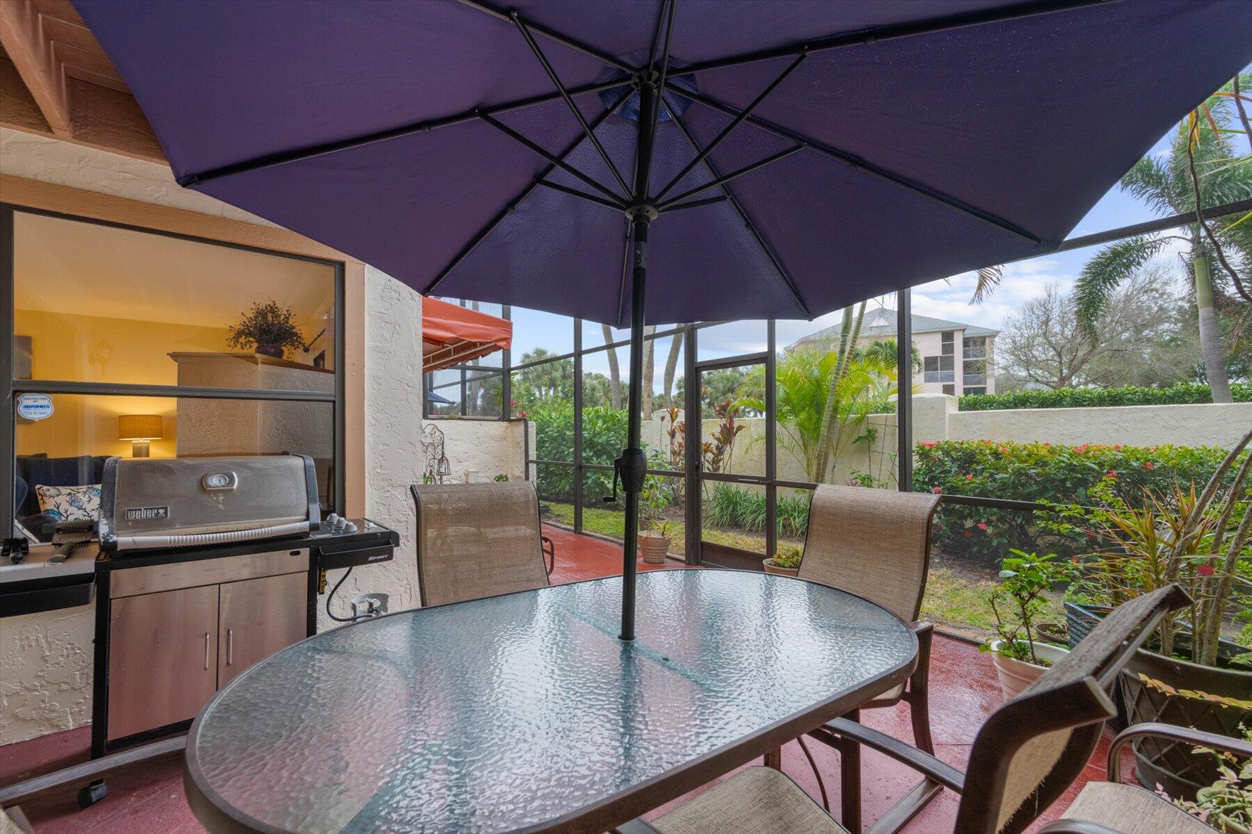 Screened patio with a glass-top dining table, large navy umbrella, Weber grill, and landscaped garden.