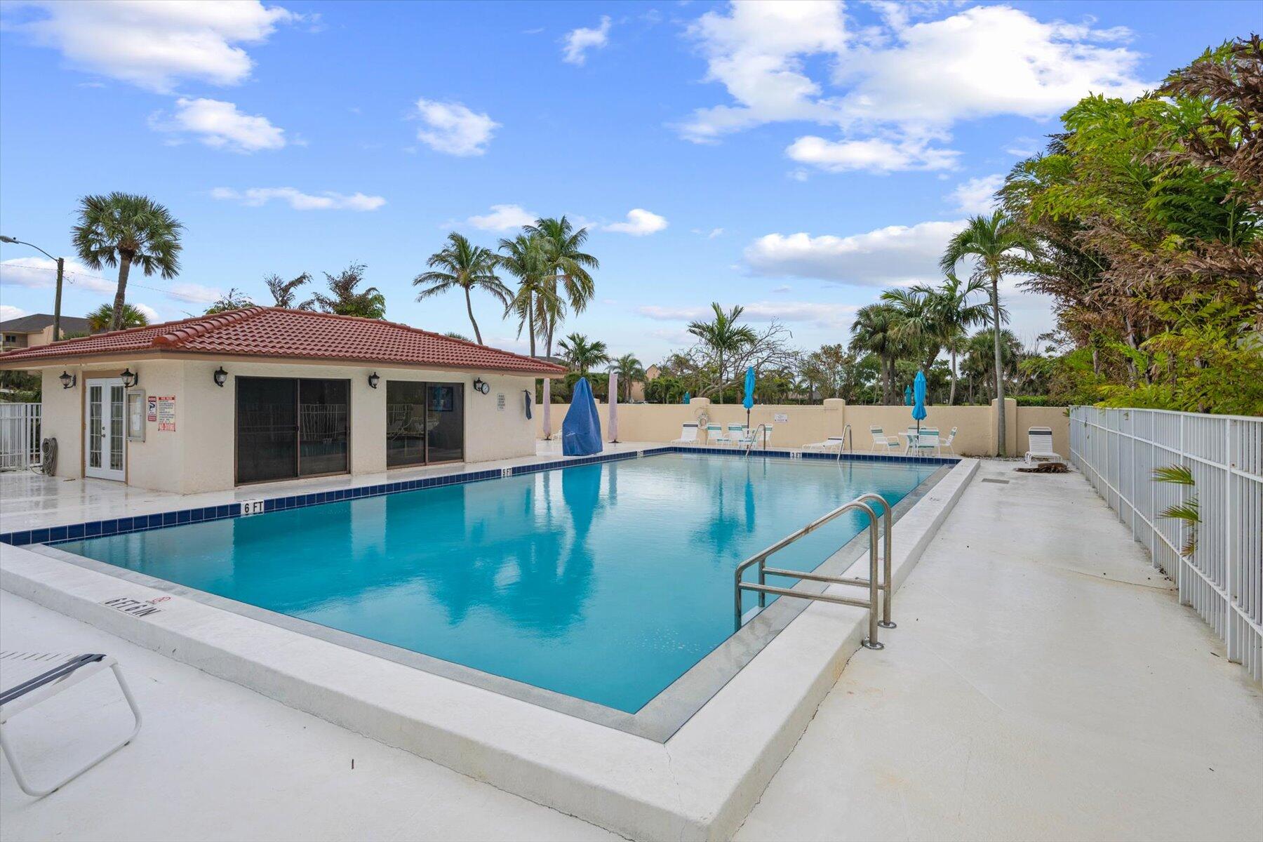 Community swimming pool beside a red-tile-roof clubhouse, with blue umbrellas and palm trees.