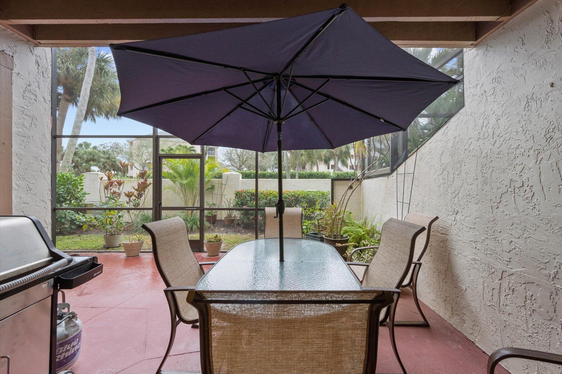 Covered patio with umbrella-shaded dining set, grill, and views of a tropical garden.
