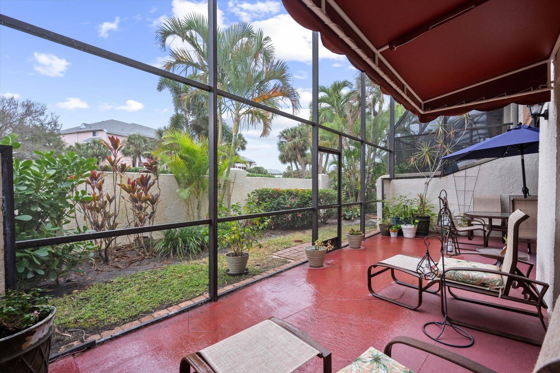 Screened patio with a burgundy awning, red floor, outdoor seating, and tropical landscaping.