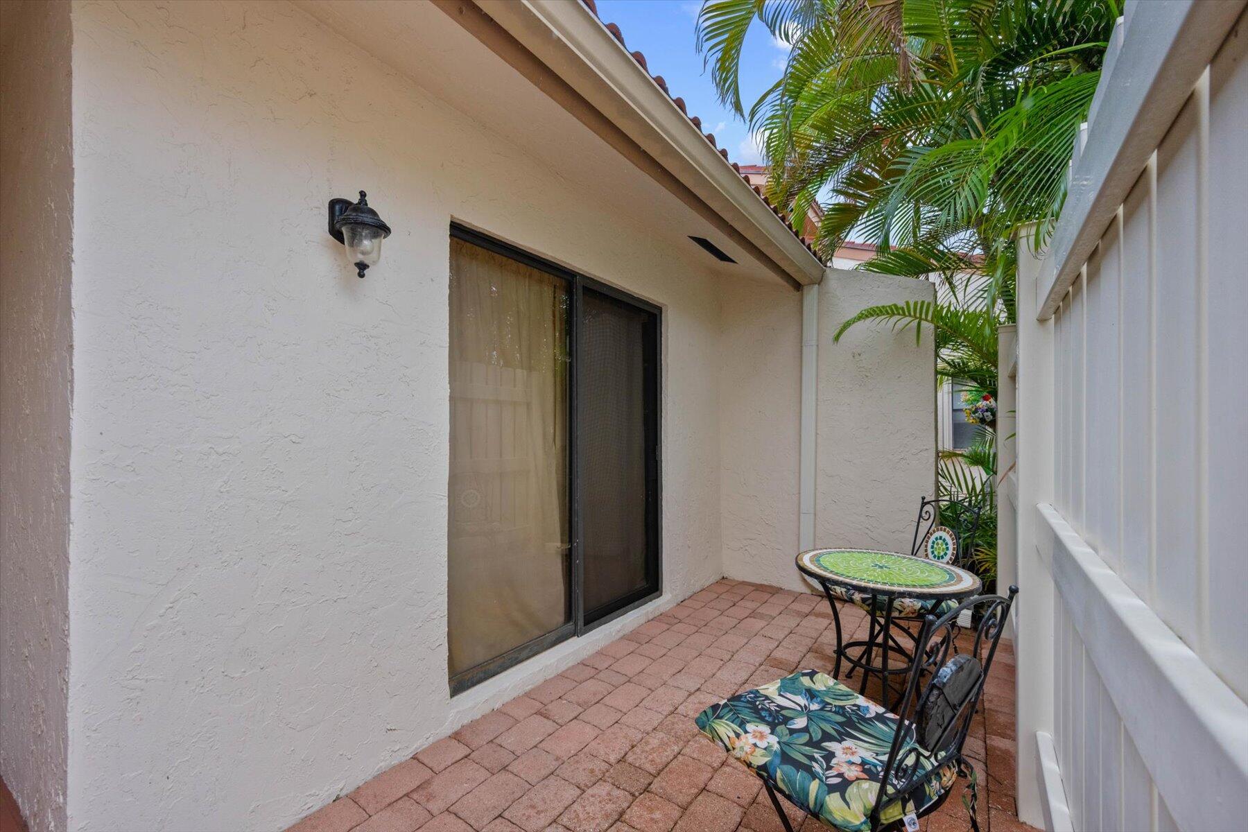 Brick patio with a mosaic table and cushioned chairs beside a sliding glass door and tropical palms.
