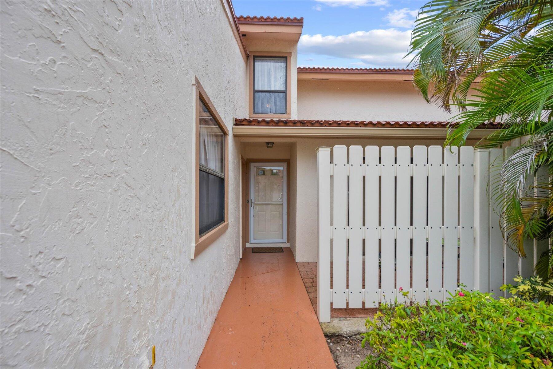 Stucco home entrance with terracotta walkway, white picket gate, and palm fronds.