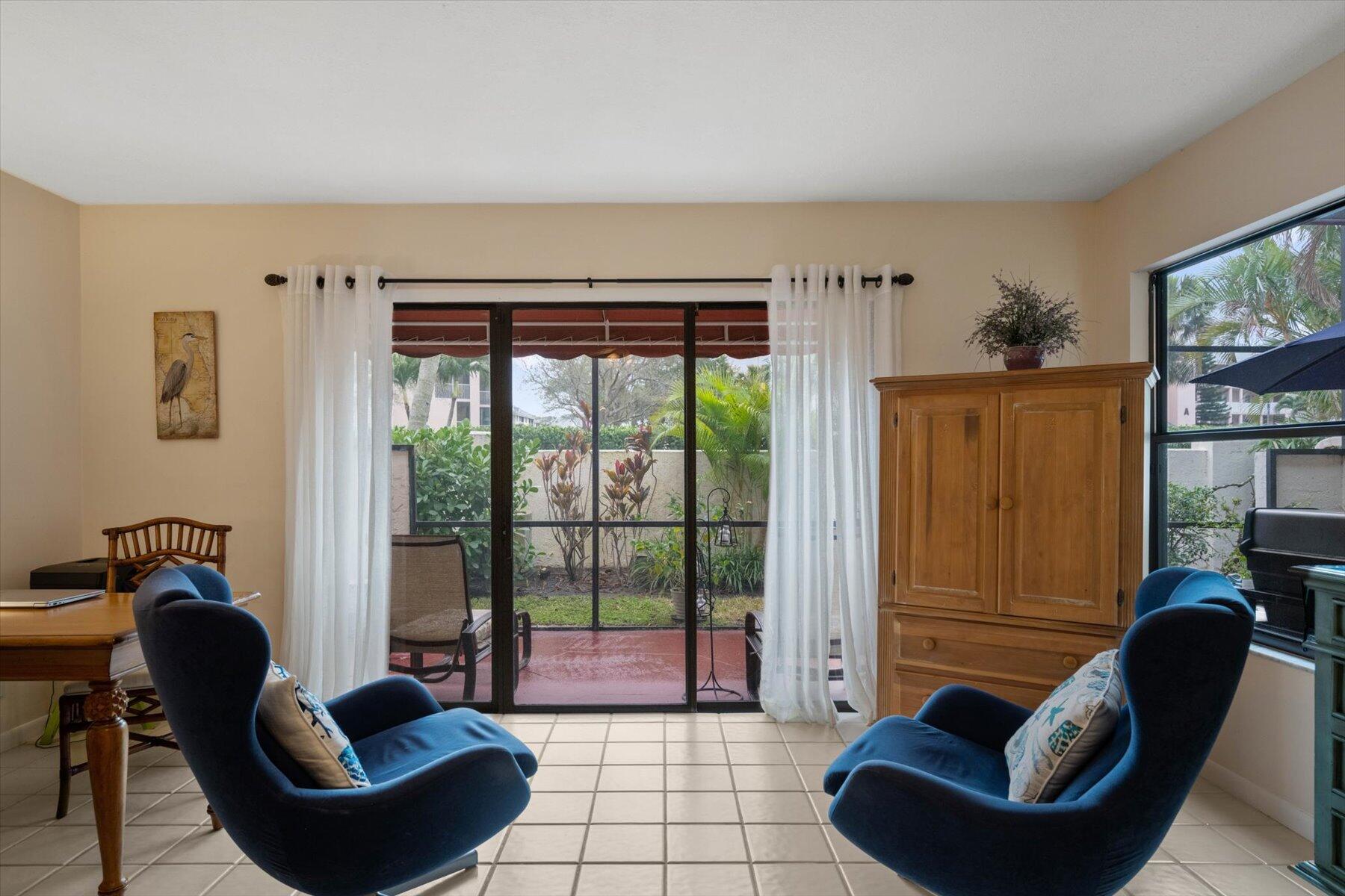 Furnished living room with blue armchairs and sliding glass doors overlooking a garden patio.