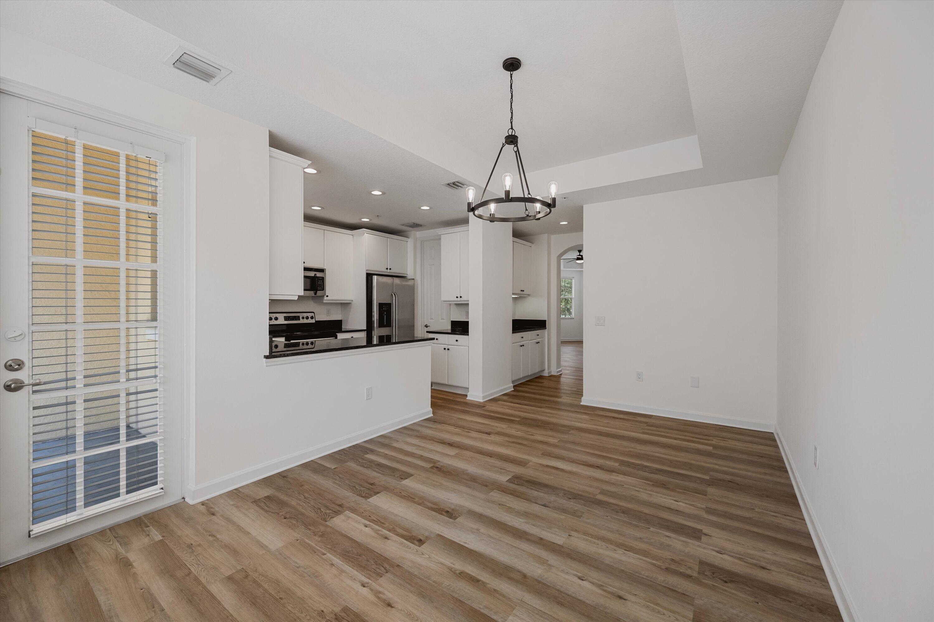 Empty dining area beside a white kitchen, with wood-look flooring and a black circular chandelier.