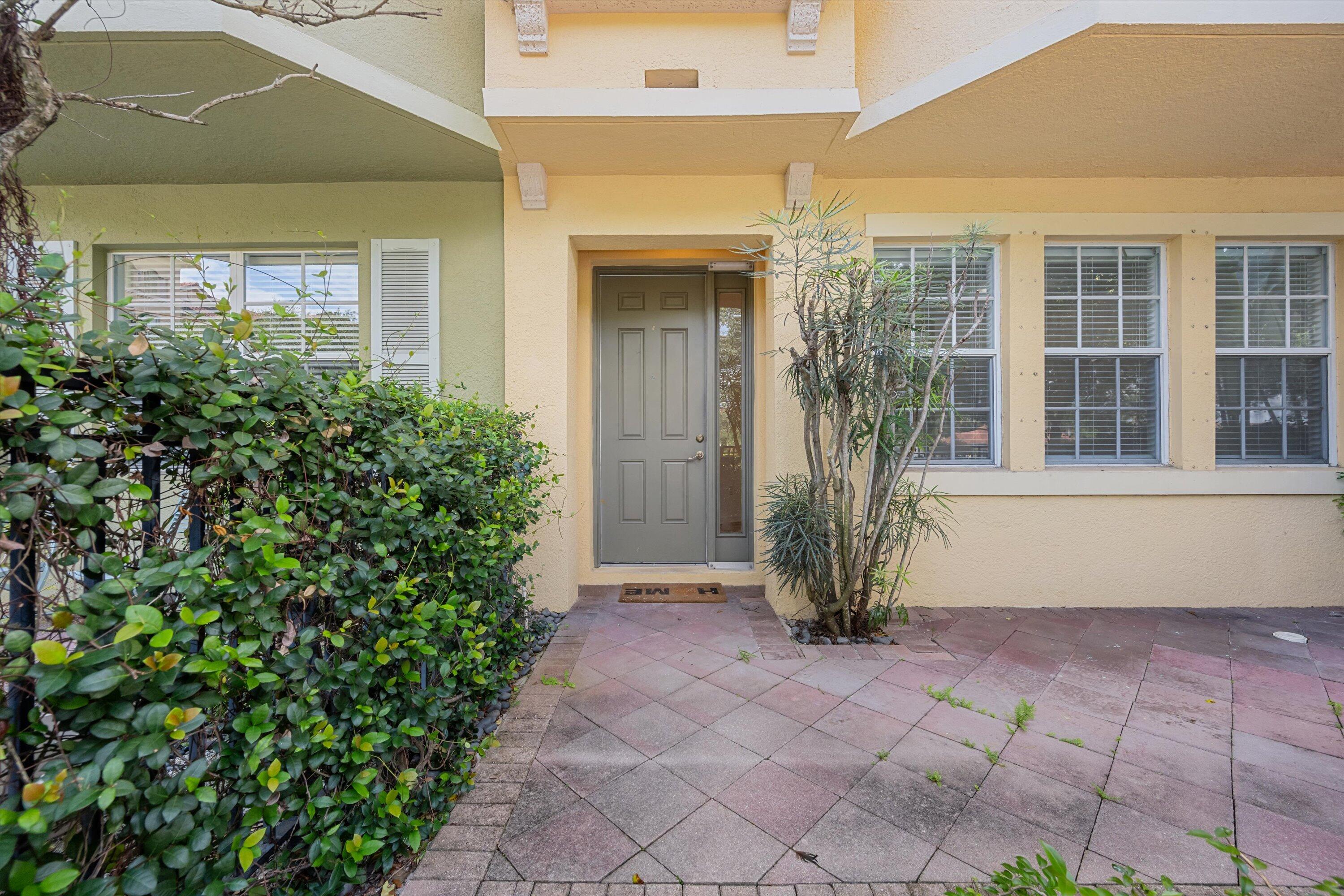 Home entrance with a gray front door, covered porch, shuttered windows, and a paved walkway bordered by shrubs.