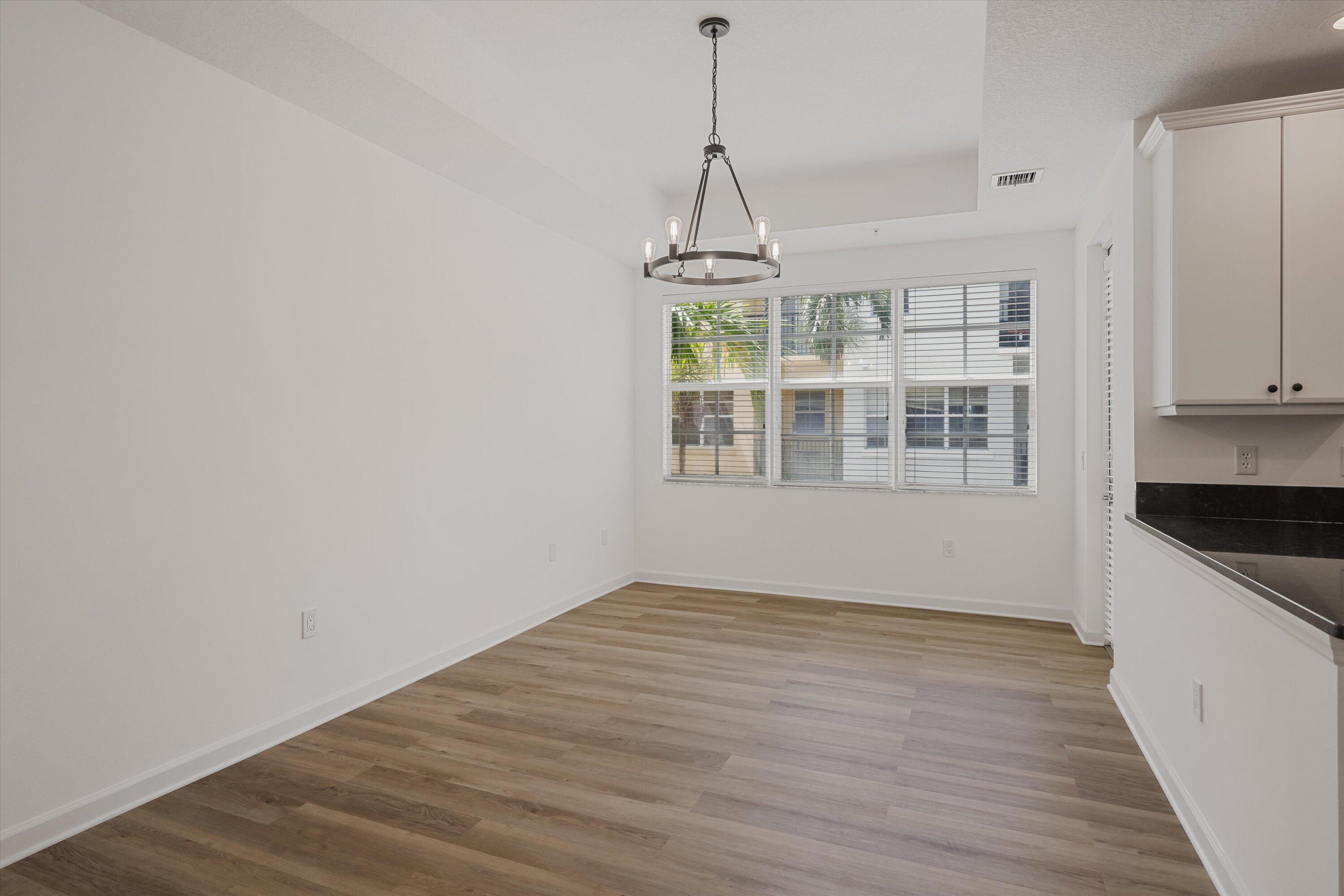 Empty dining area with wood-look floors, a metal chandelier, wide window, and adjoining kitchen counter.