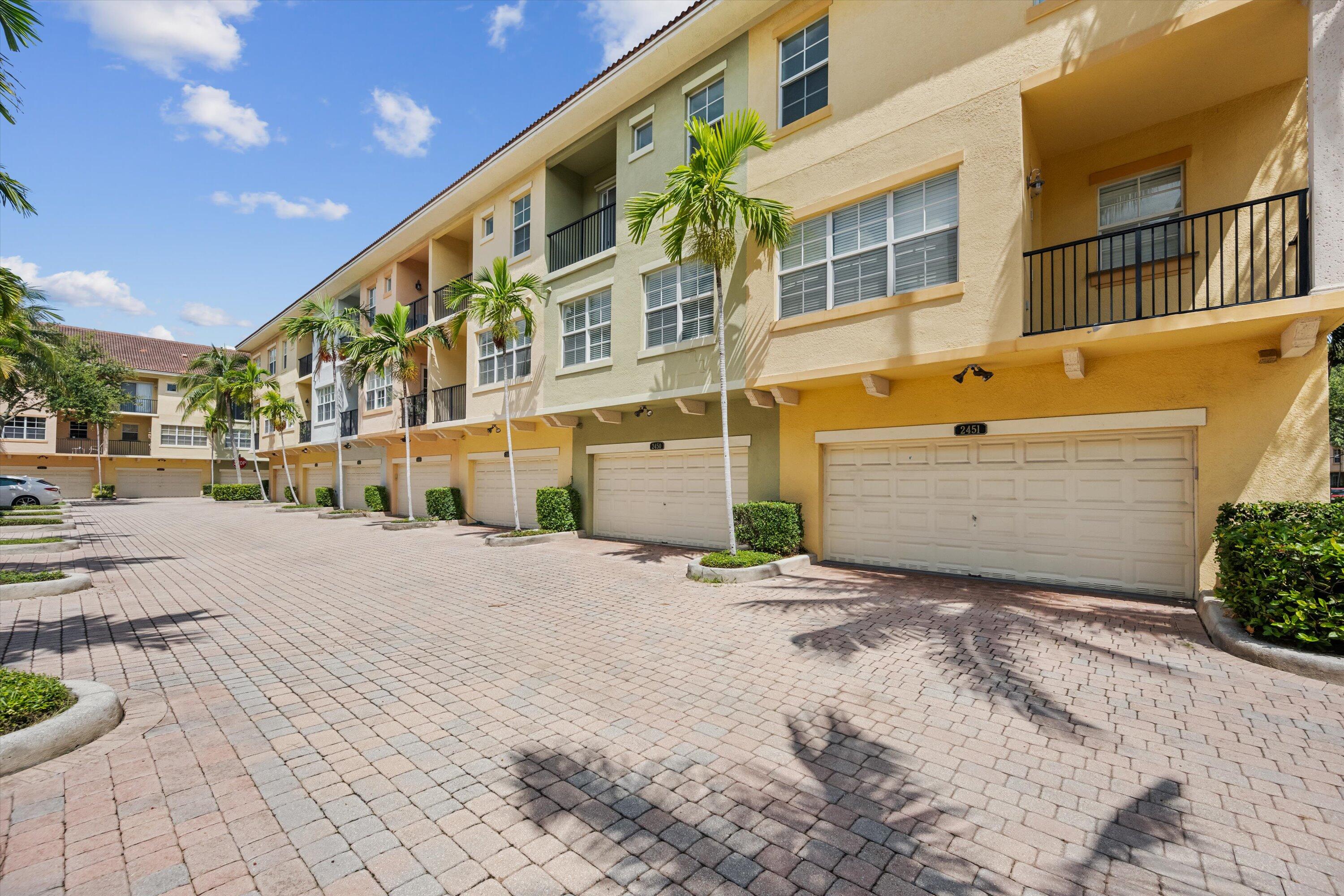 Attached residences with balconies and garage doors along a palm-lined brick driveway.