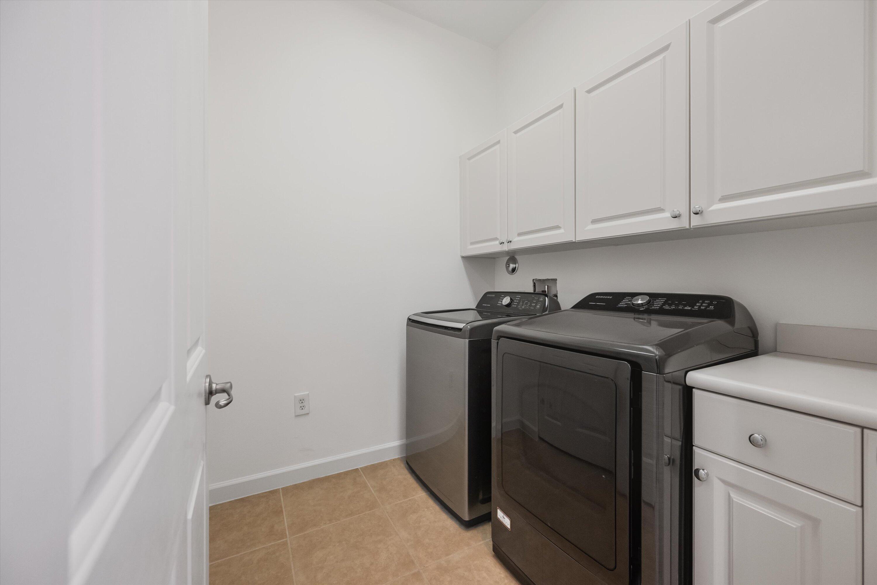 Laundry room with dark washer and dryer, white overhead cabinets, and tan tile floor.