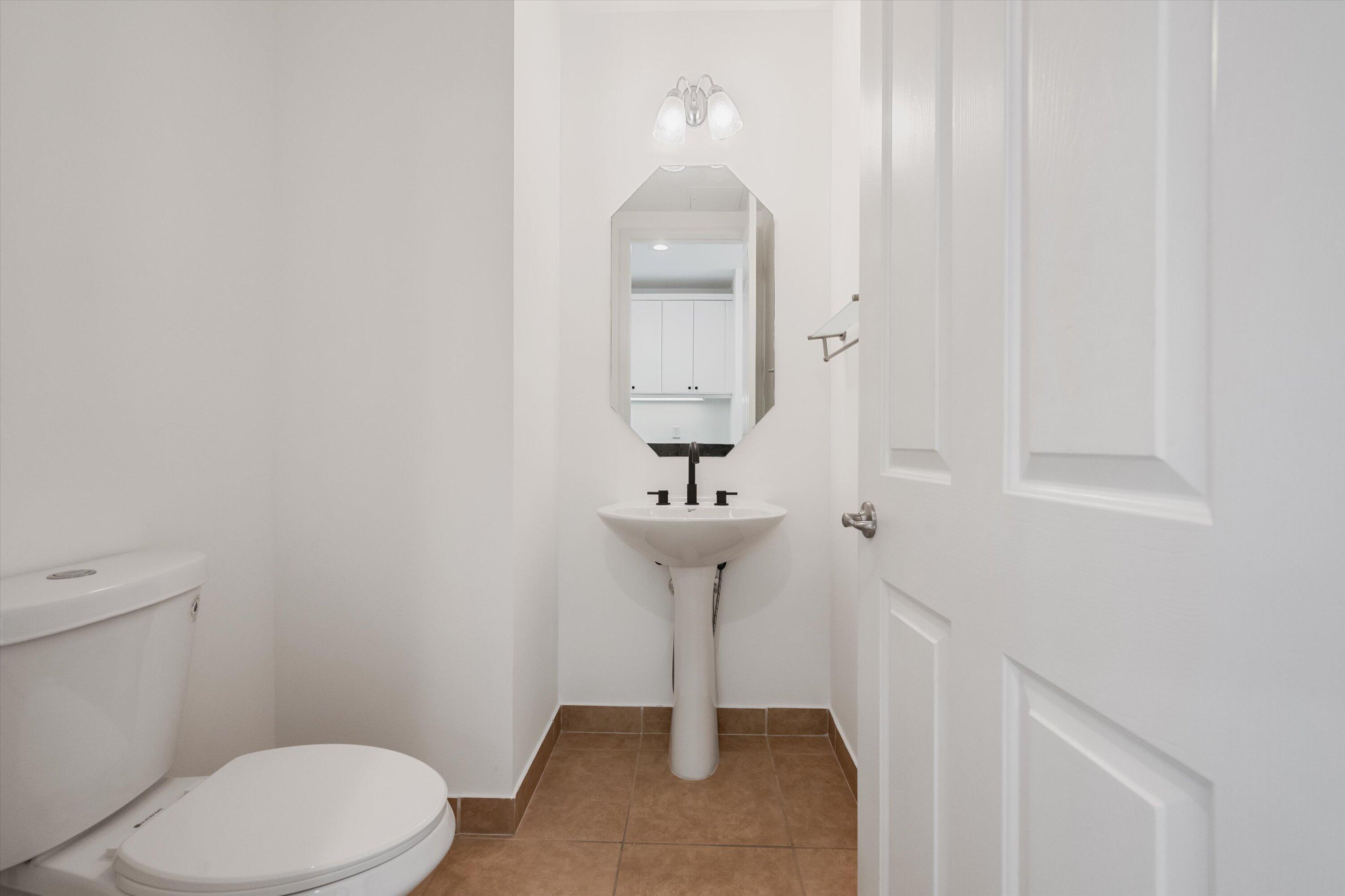 Bathroom with a white toilet, pedestal sink, black fixtures, octagonal mirror, and brown tile floor.